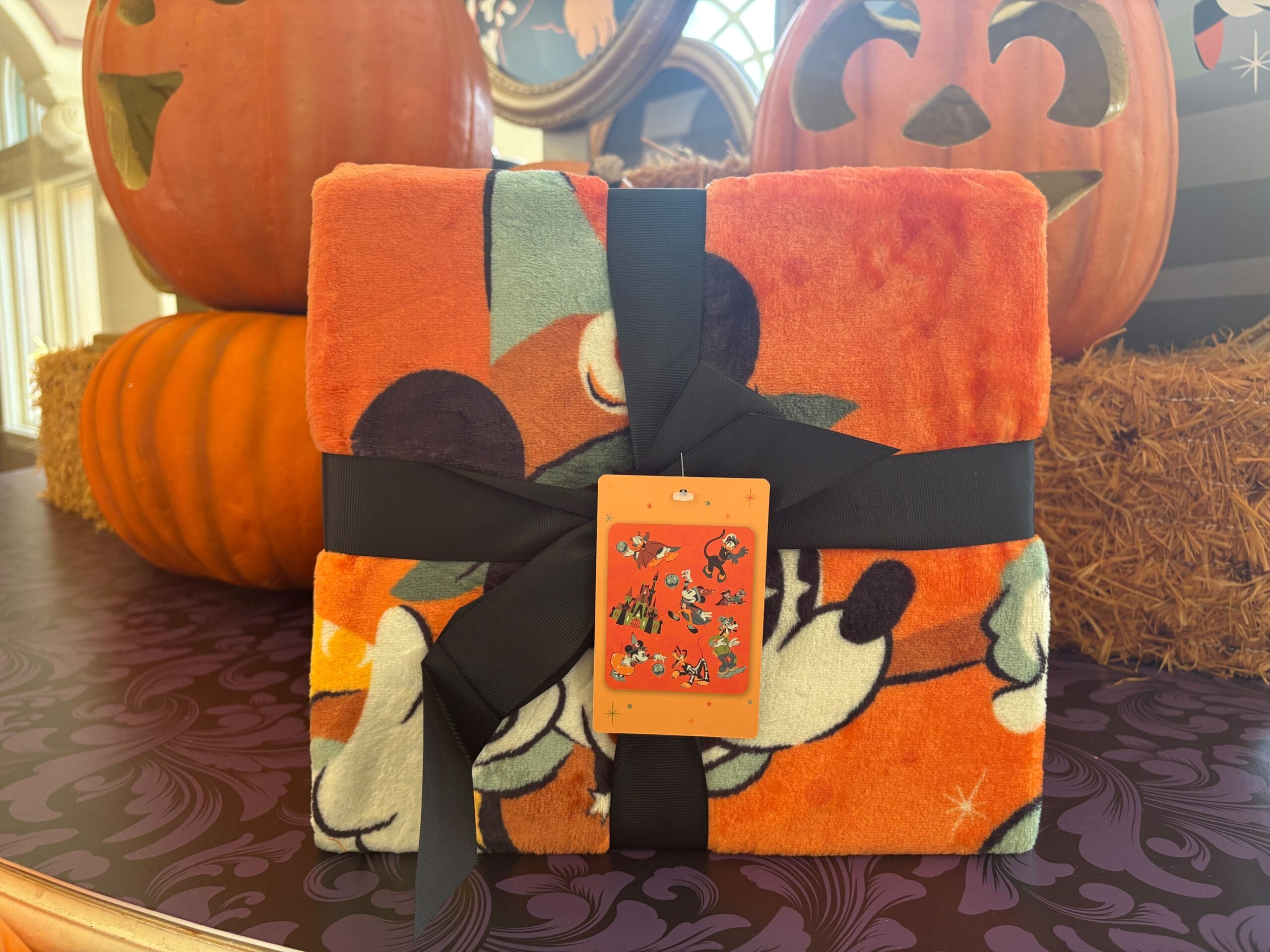 An orange Mickey Mouse blanket with a black ribbon sits among Walt Disney World Halloween merchandise and pumpkins.