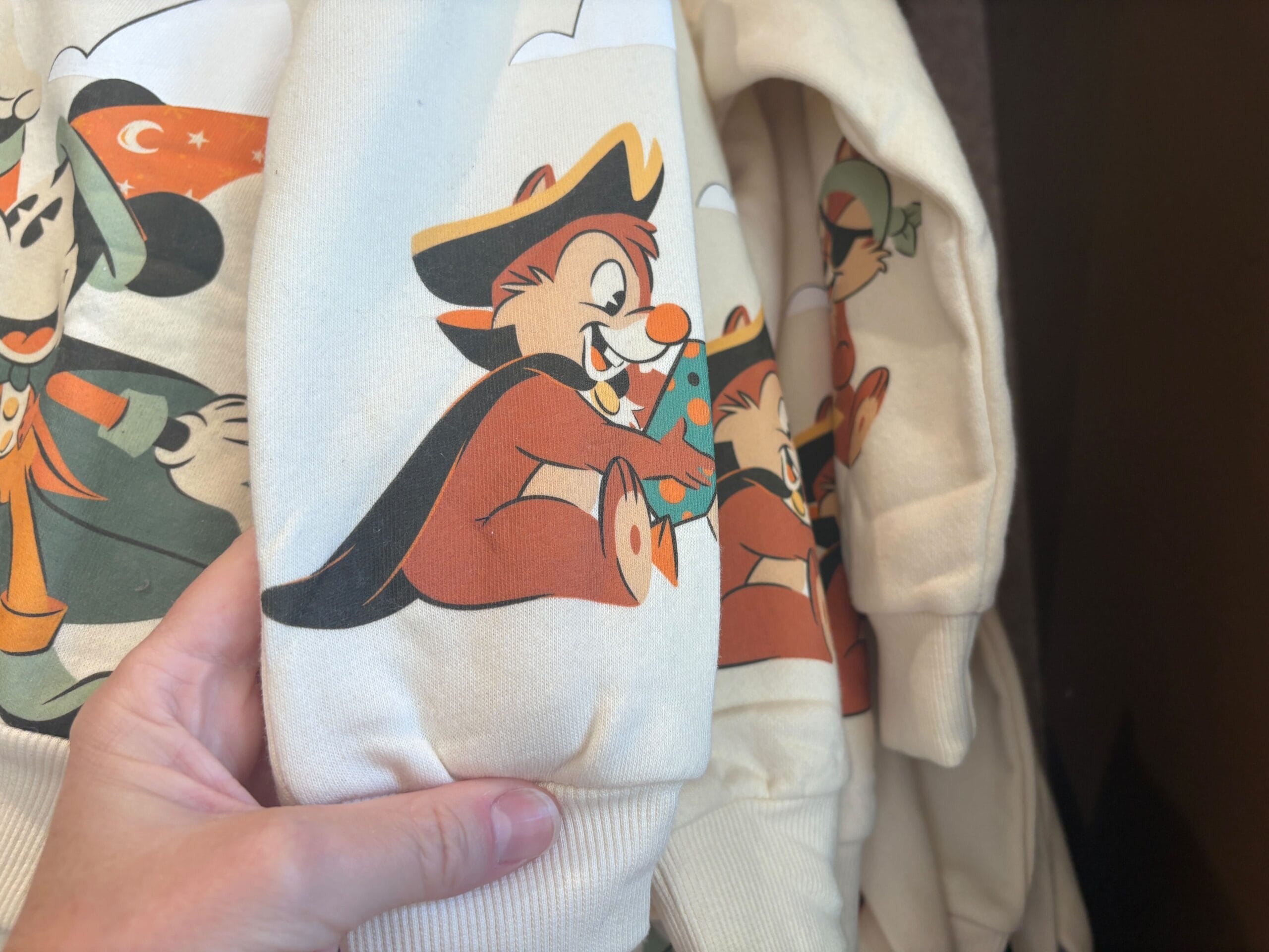 A hand holds a cream-colored sweater, part of Walt Disney World Halloween merchandise, with a chipmunk in a hat and tree.