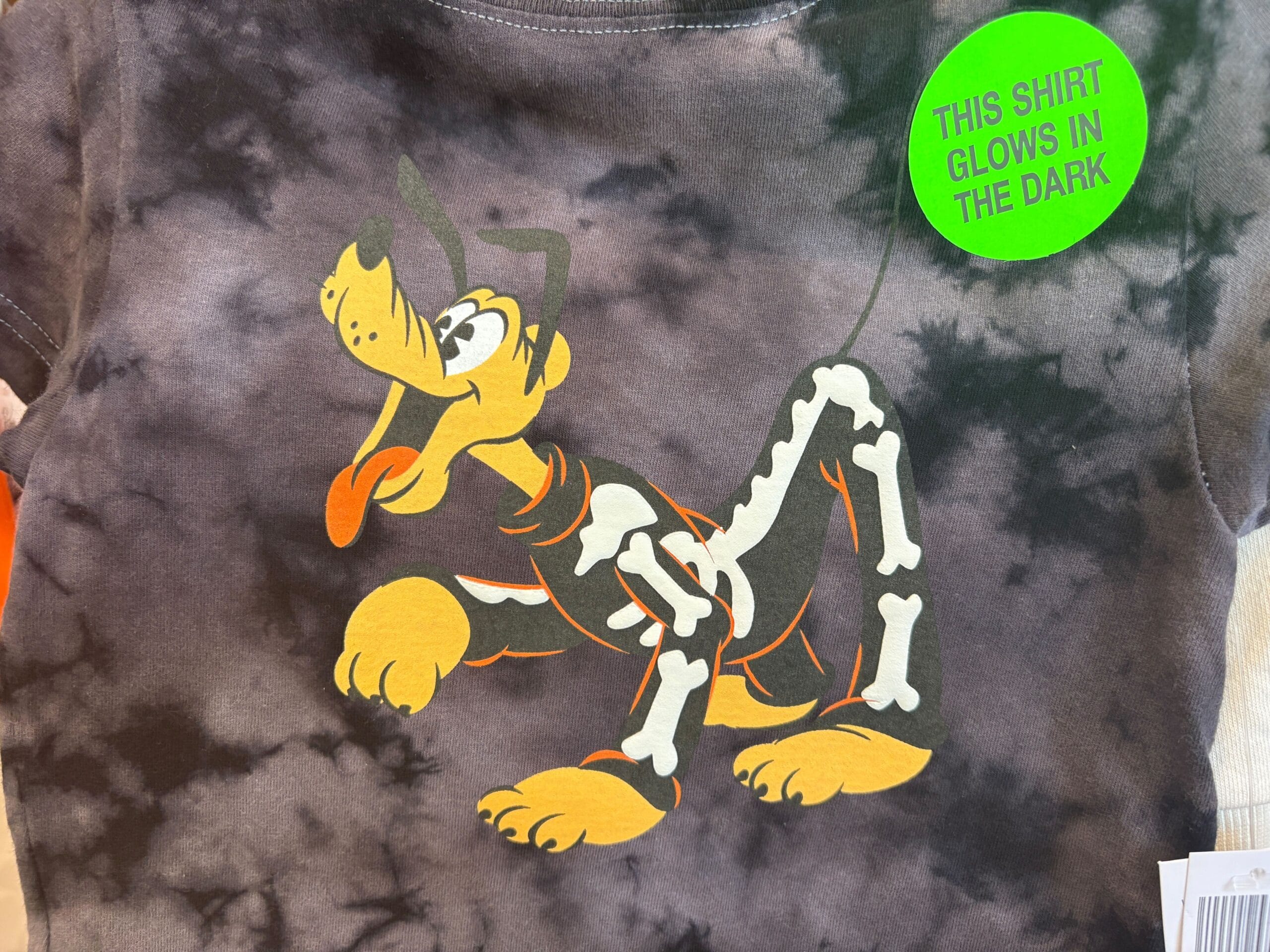 Gray tie-dye shirt featuring Pluto and bones, with a sticker—part of Walt Disney World Halloween Merchandise that glows in the dark.