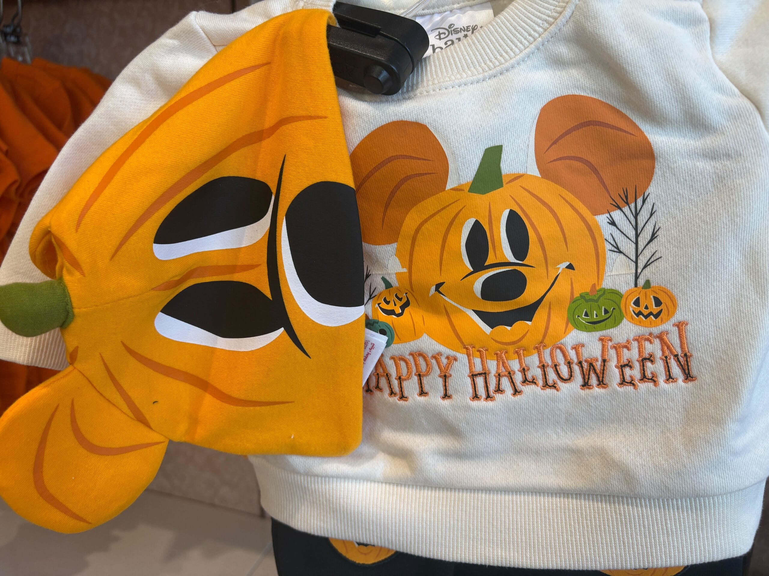 White sweatshirt with Walt Disney World pumpkin Mickey Mouse and "Happy Halloween" text, next to an orange pumpkin hat.