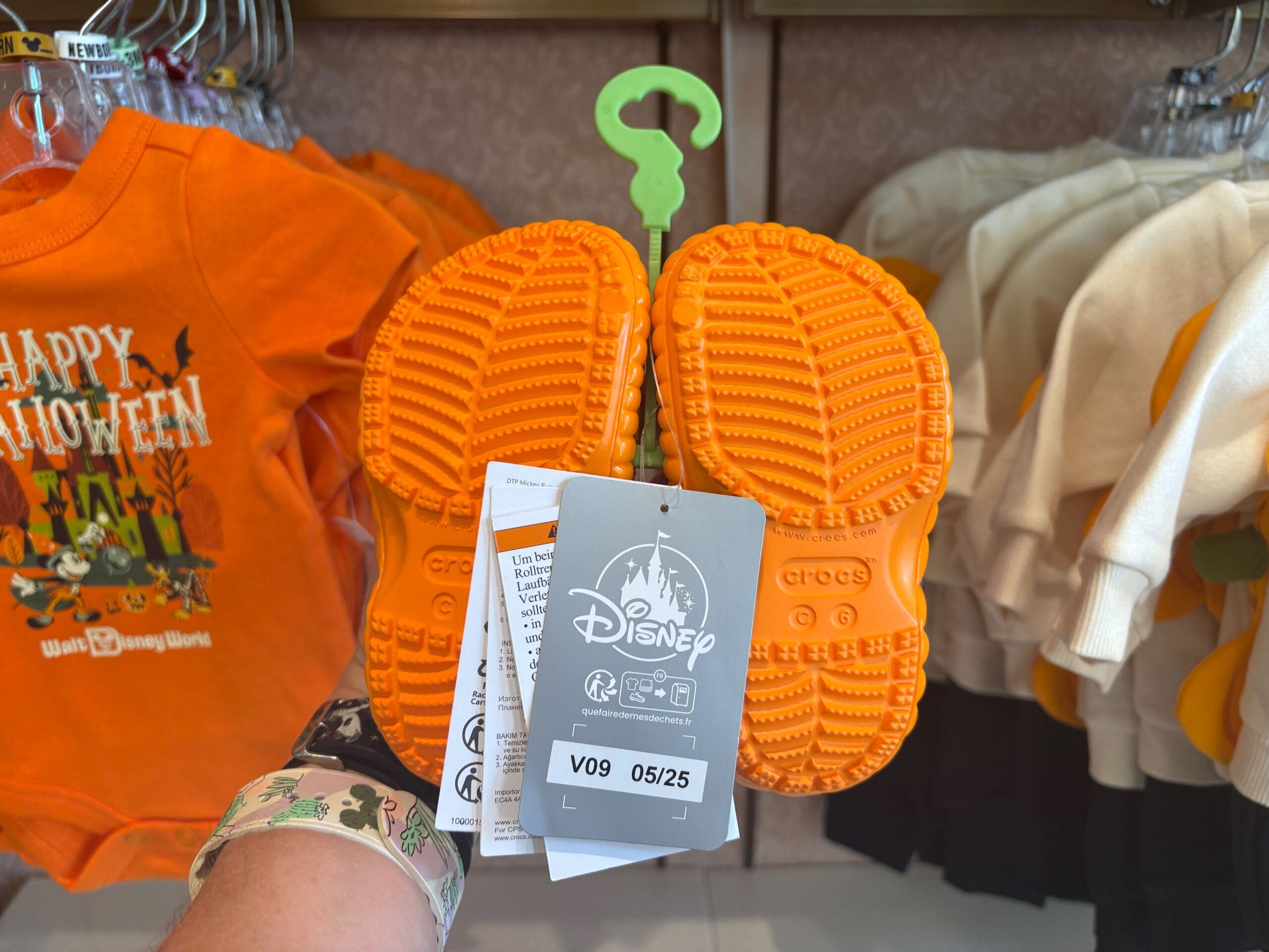A hand holds orange Crocs with Disney tags in a store, surrounded by Walt Disney World Halloween merchandise.