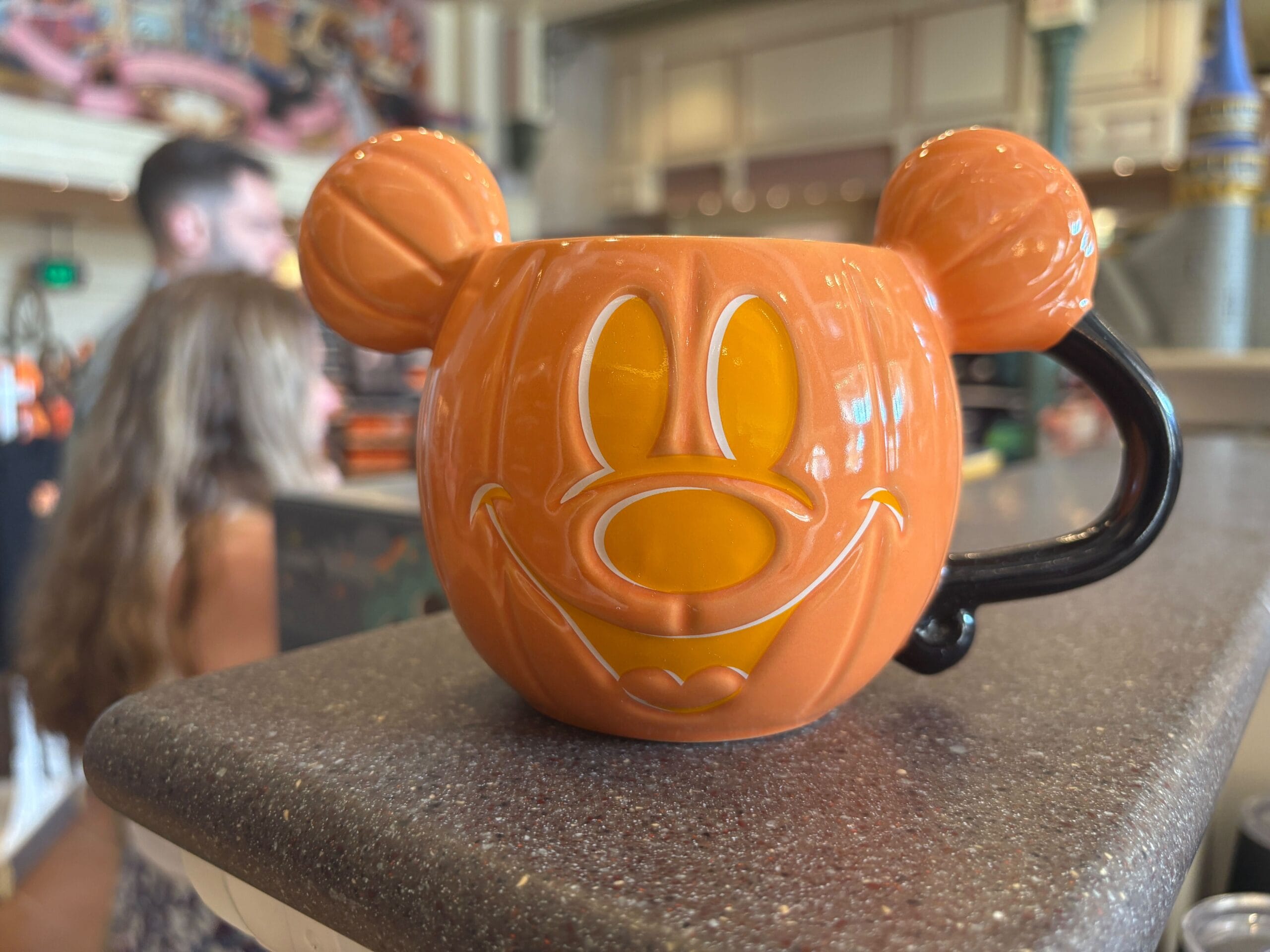 An orange mug shaped like a Mickey Mouse pumpkin sits on a countertop with other Walt Disney World Halloween merchandise.