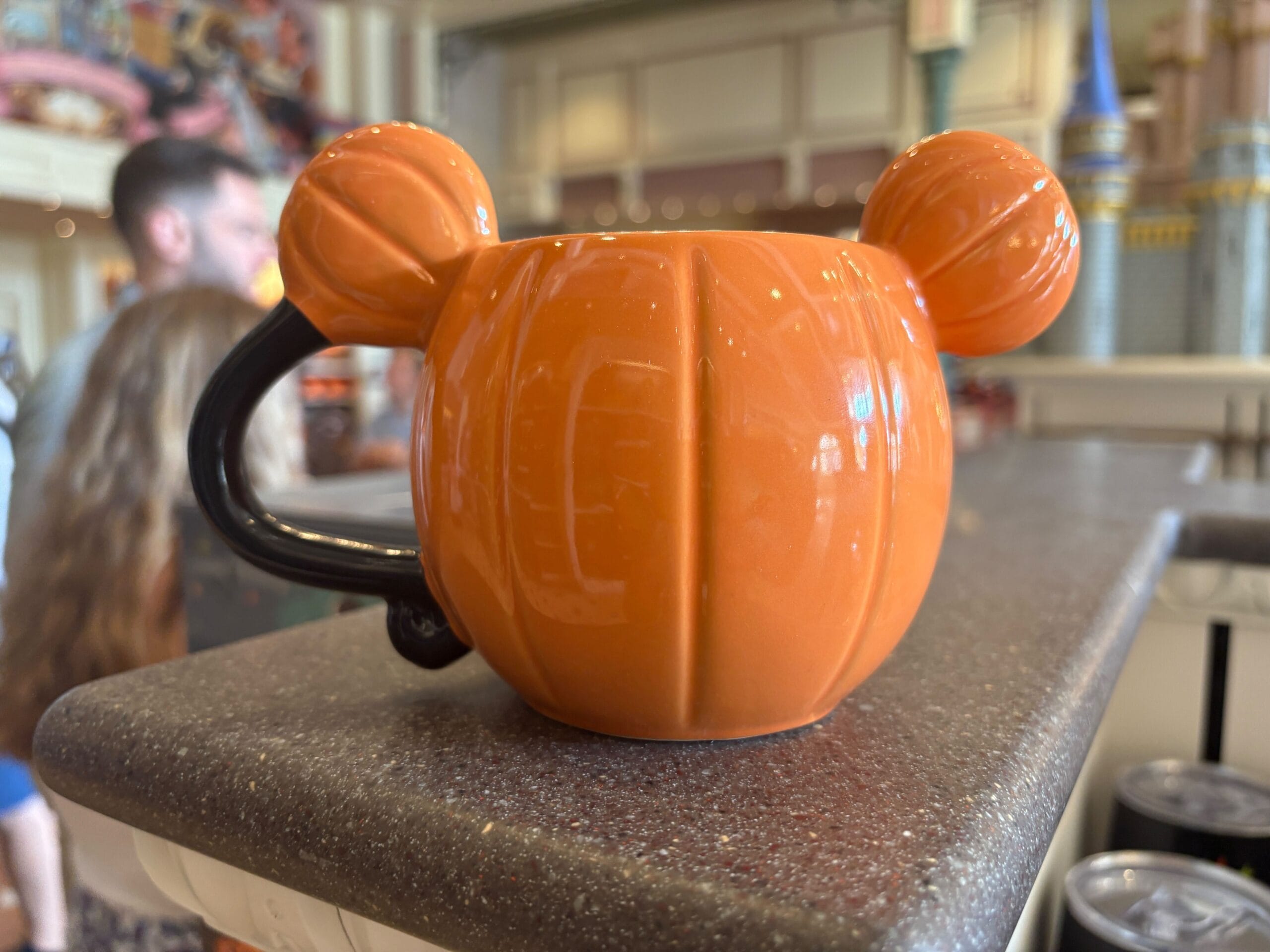 An orange pumpkin mug with Mickey Mouse ears, part of Walt Disney World Halloween Merchandise, sits on a shop countertop.