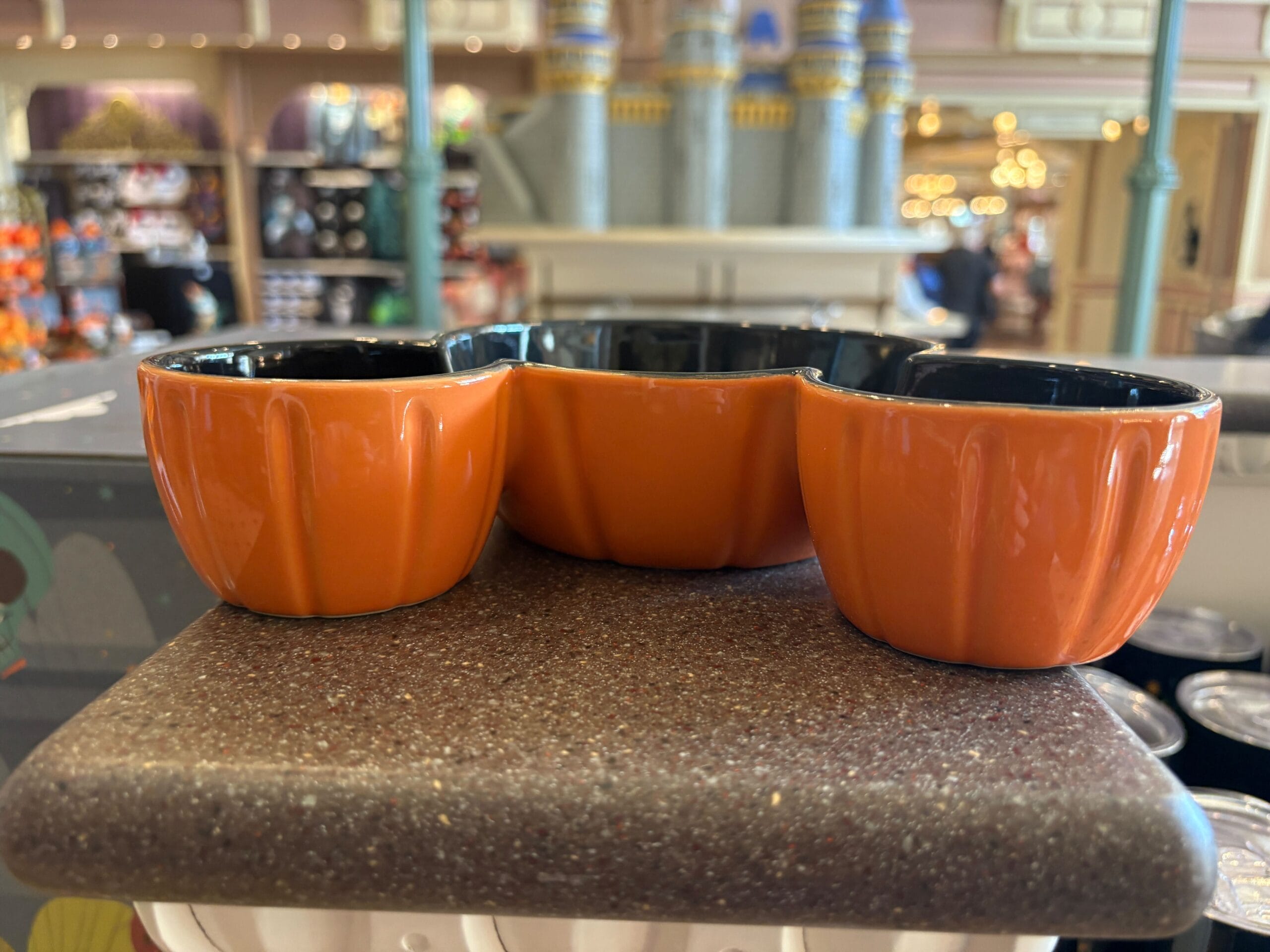 Three orange pumpkin-shaped ceramic bowls joined together, part of Halloween merchandise, displayed on a brown shelf.