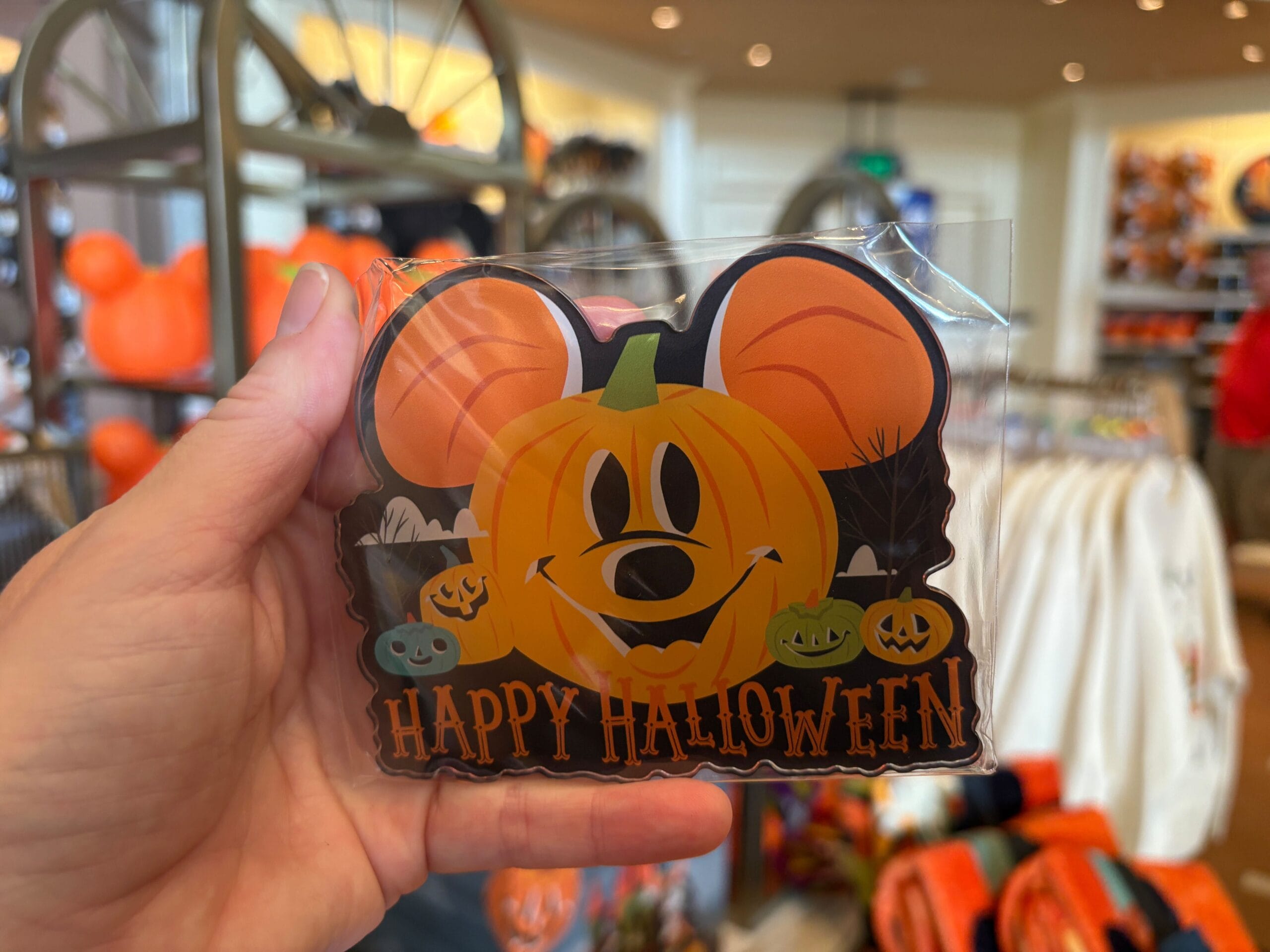 Hand holding a "Happy Halloween" pumpkin sign with Mickey ears, part of Walt Disney World Halloween merchandise in store.