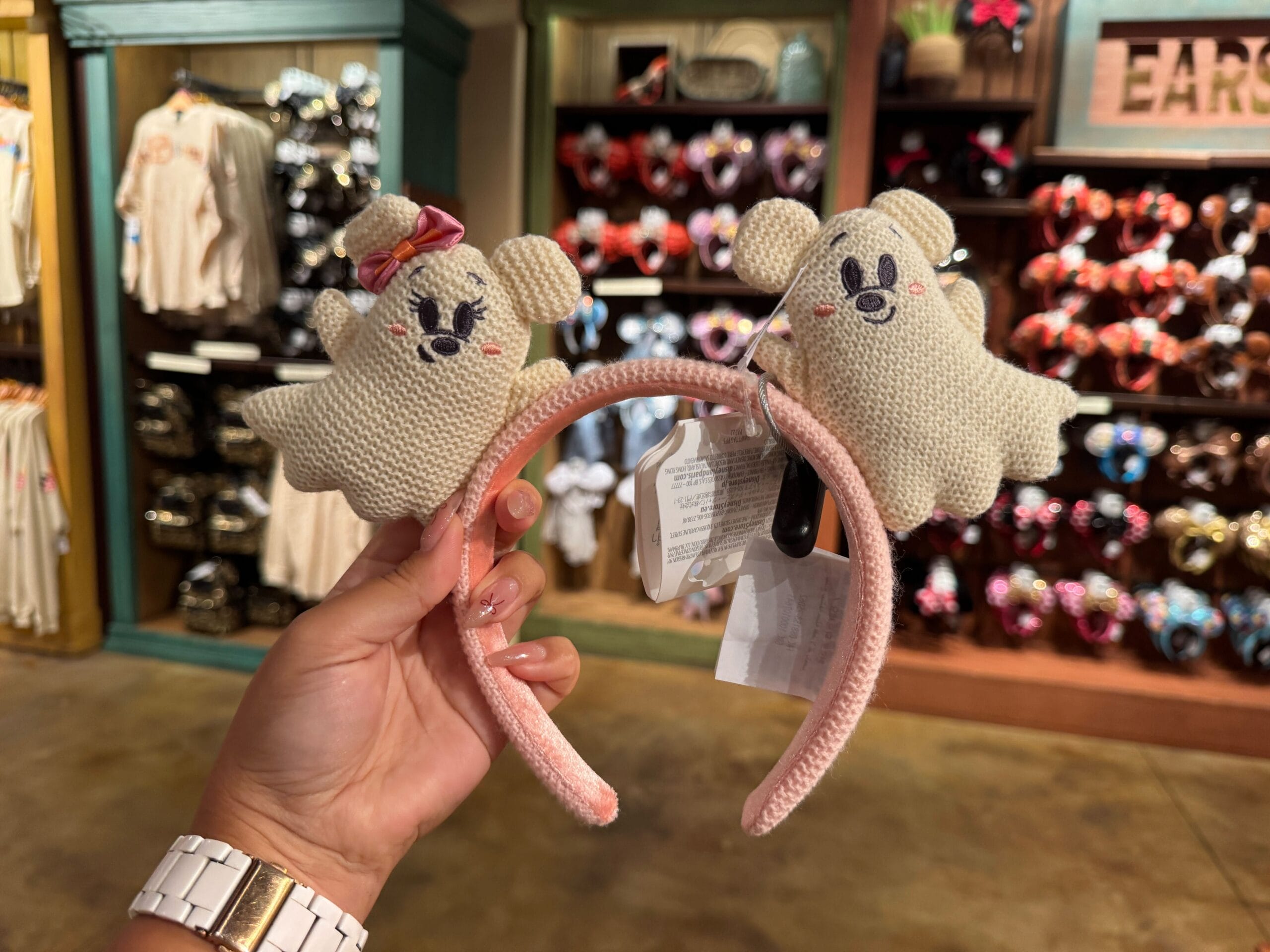 A hand holds a pink headband with two plush ghost characters among Walt Disney World Halloween merchandise.