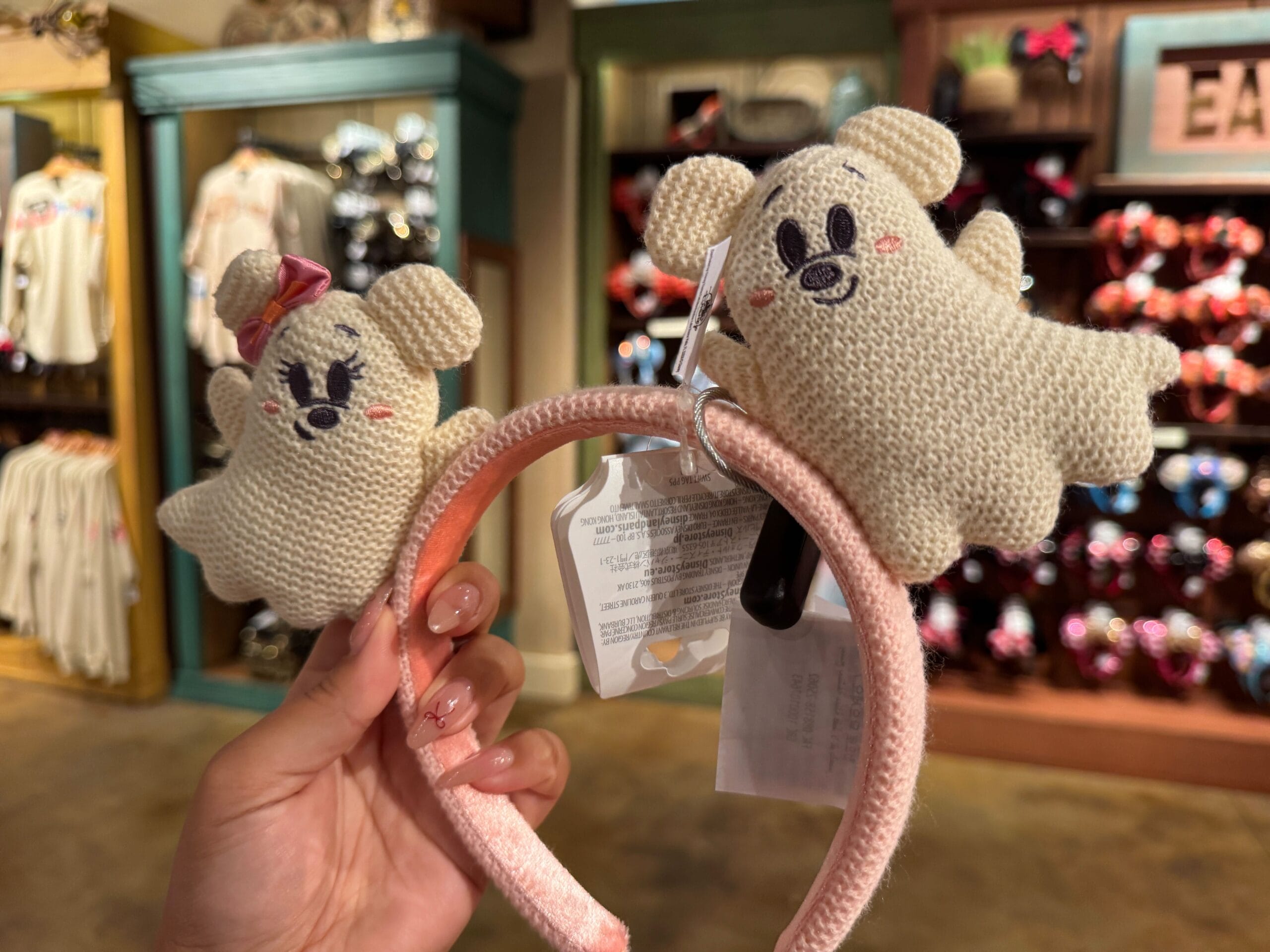 A hand holds a pink headband with two knit ghost decorations, part of the Halloween merchandise at Walt Disney World.