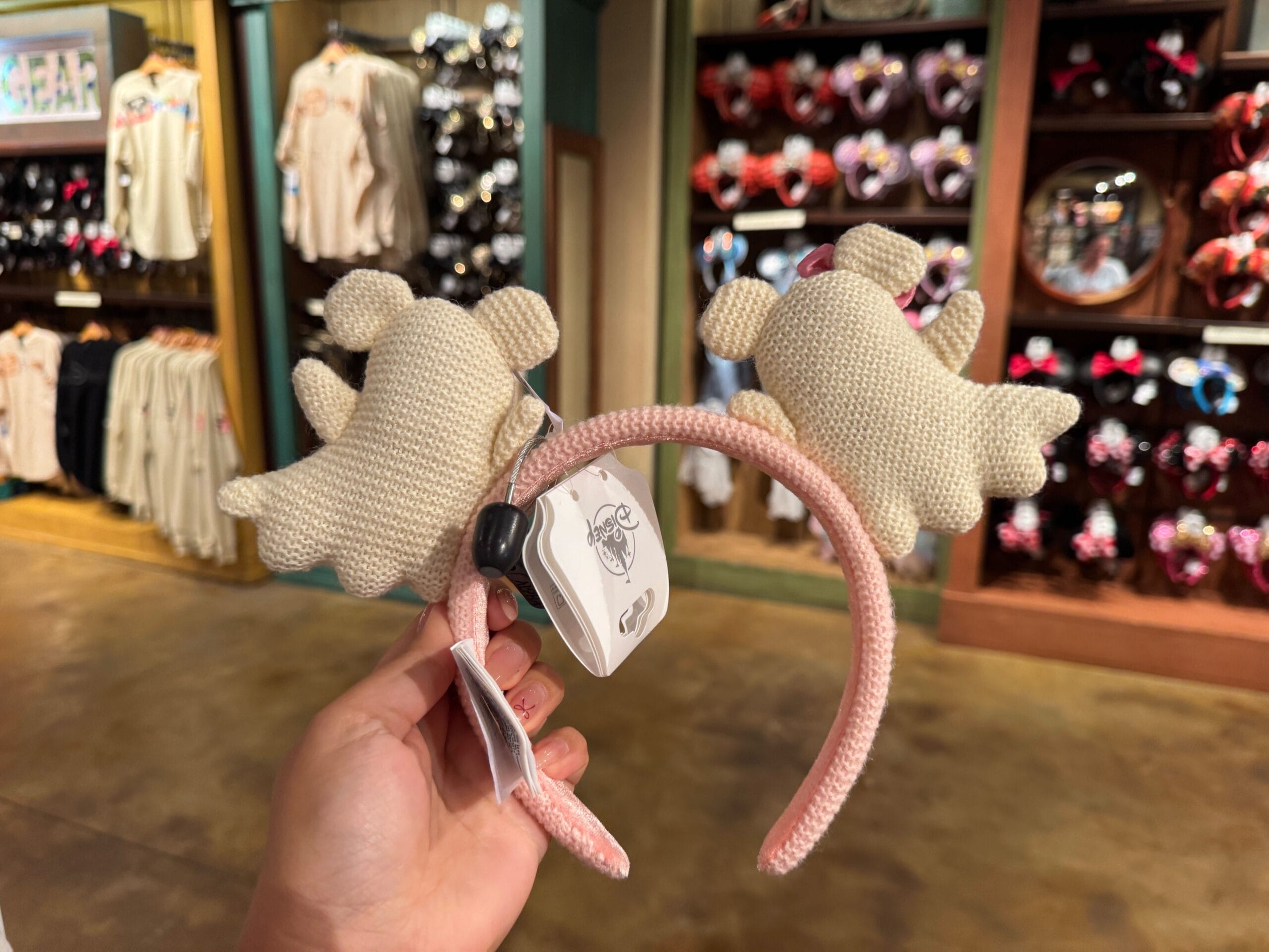 A hand holds a headband with two plush ghost shapes in a store selling Walt Disney World Halloween merchandise.