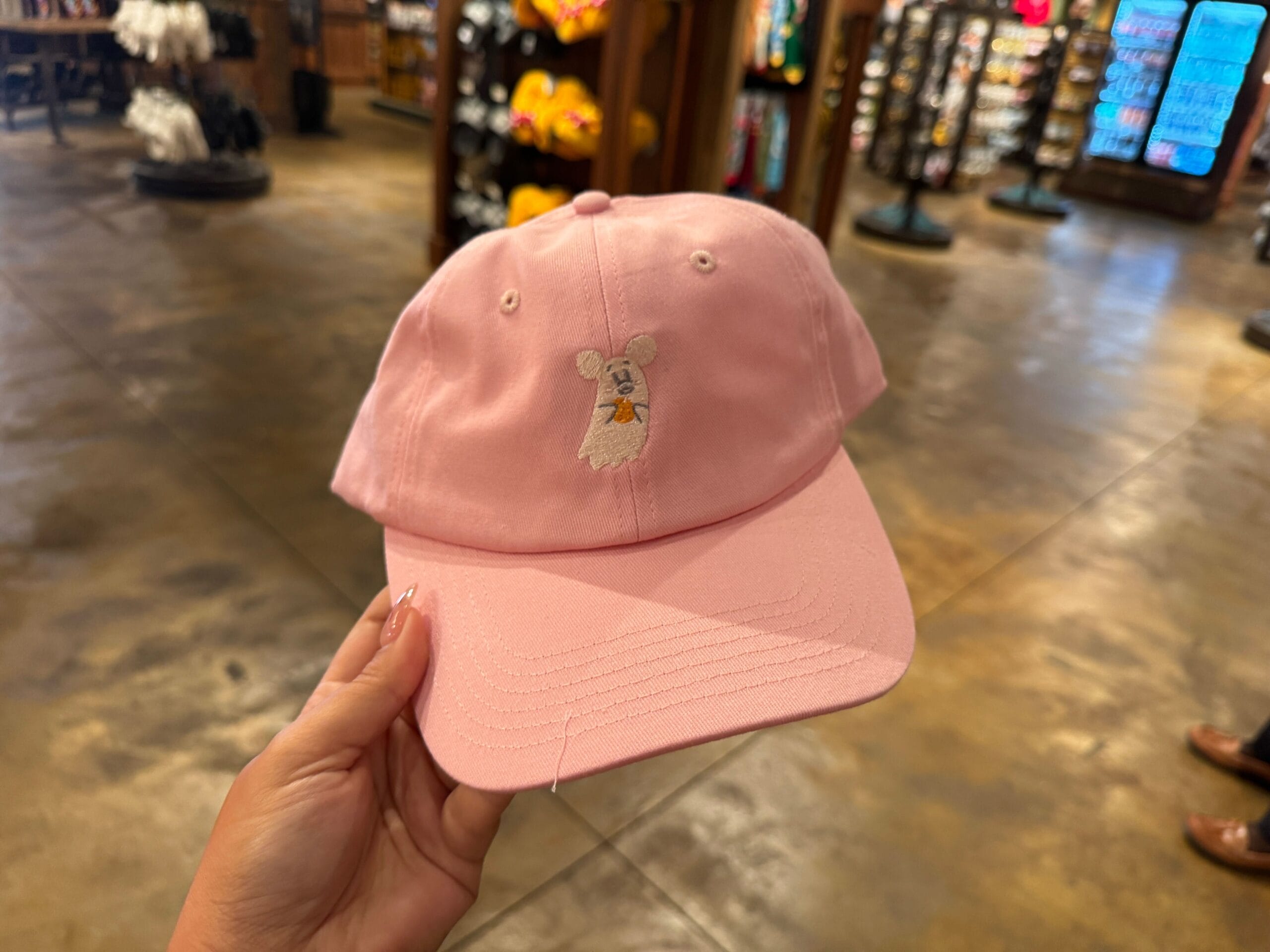 A hand holds a pink baseball cap with a cartoon mouse face, spotted among Halloween merchandise at Walt Disney World.