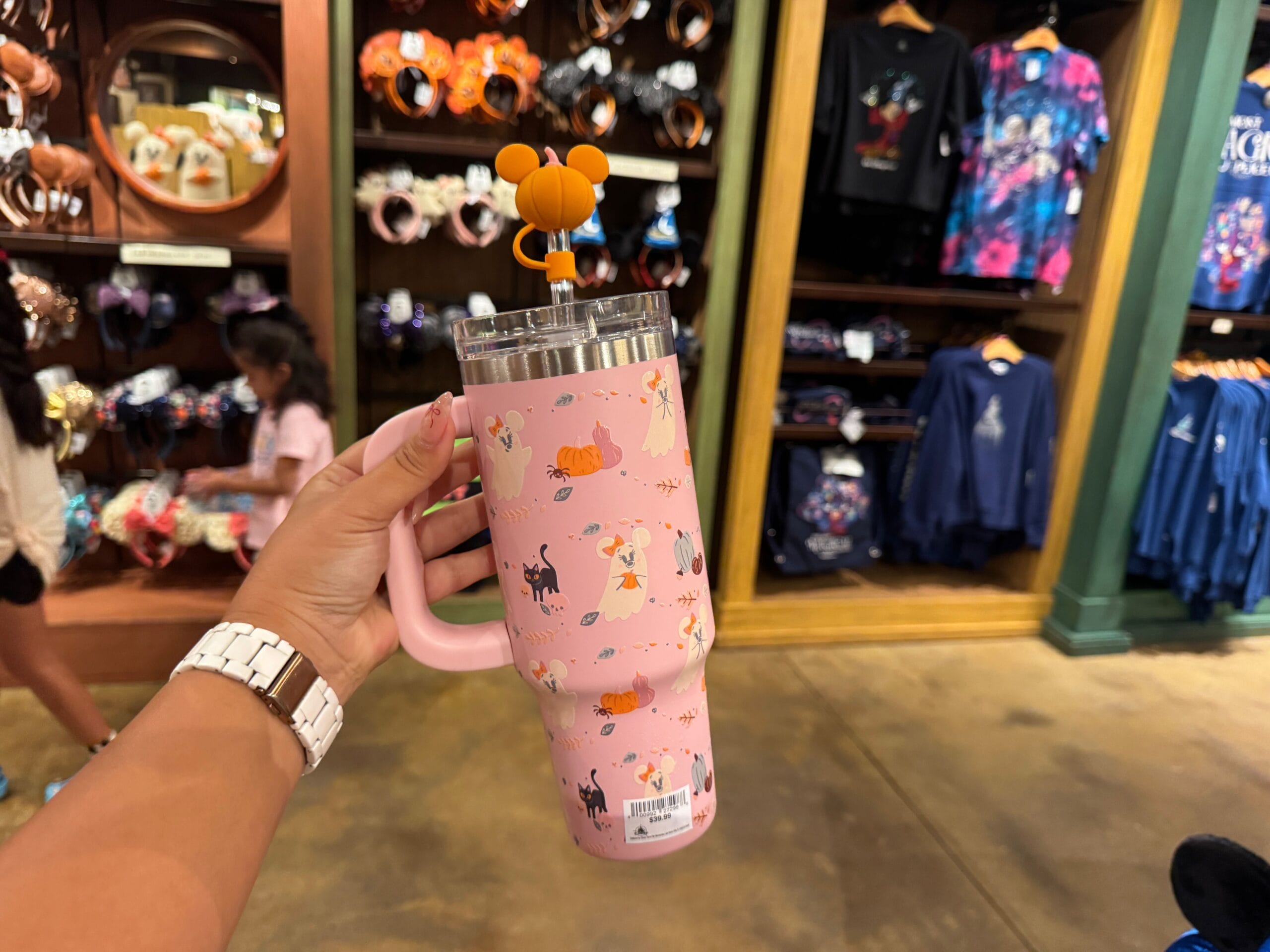 A hand holds a pink Disney princess tumbler with a Mickey straw topper among Walt Disney World clothing and accessories.