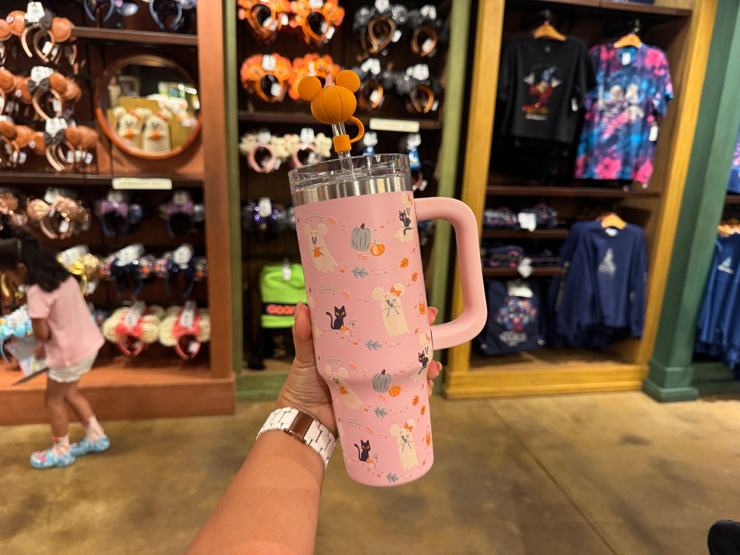 Hand holding a pink Walt Disney World Halloween tumbler in a store, with festive merchandise and clothing in the background.