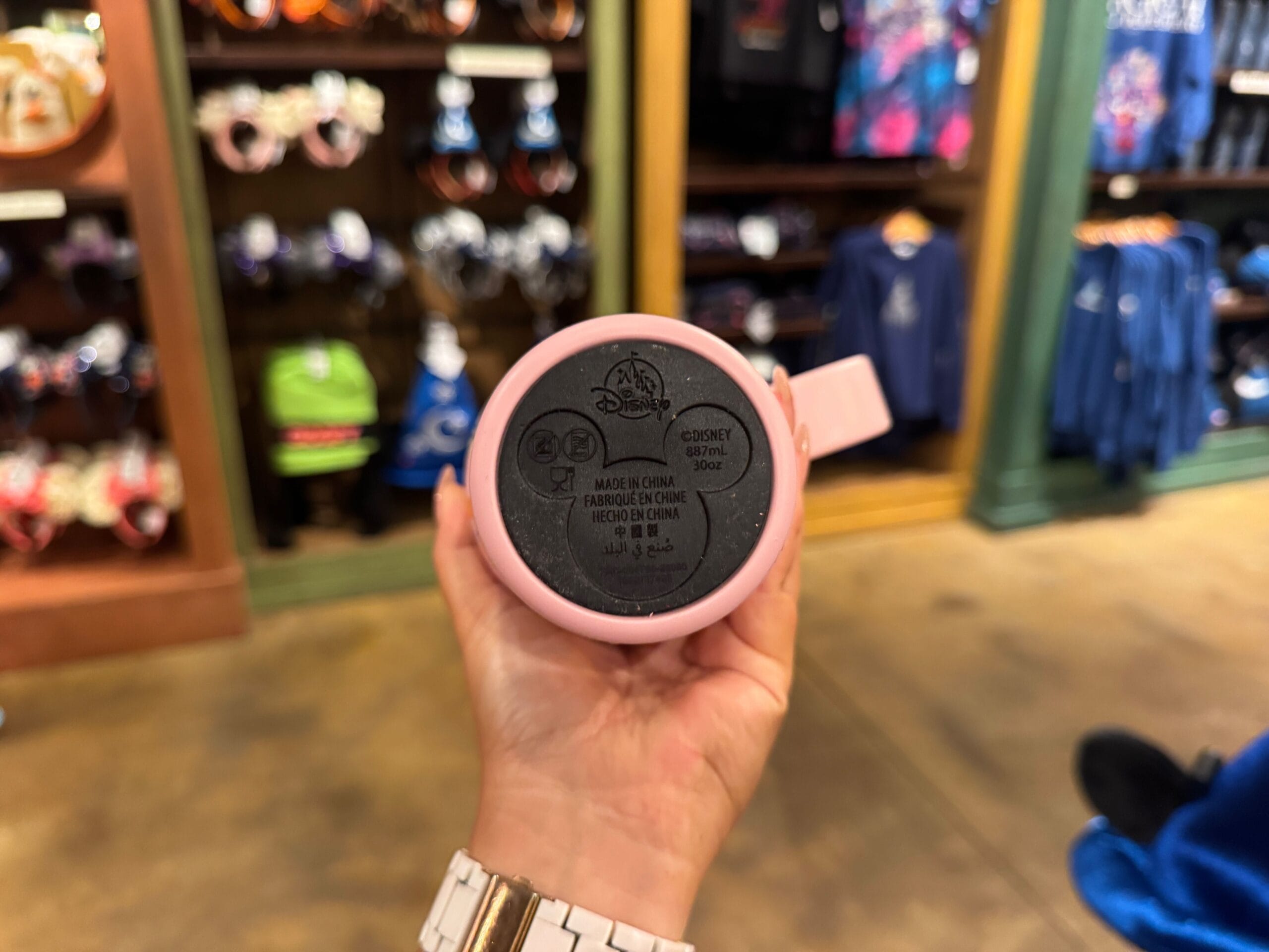 A hand holds a pink Disney mug in a store, Halloween merchandise and prices visible on shelves in the blurred background.