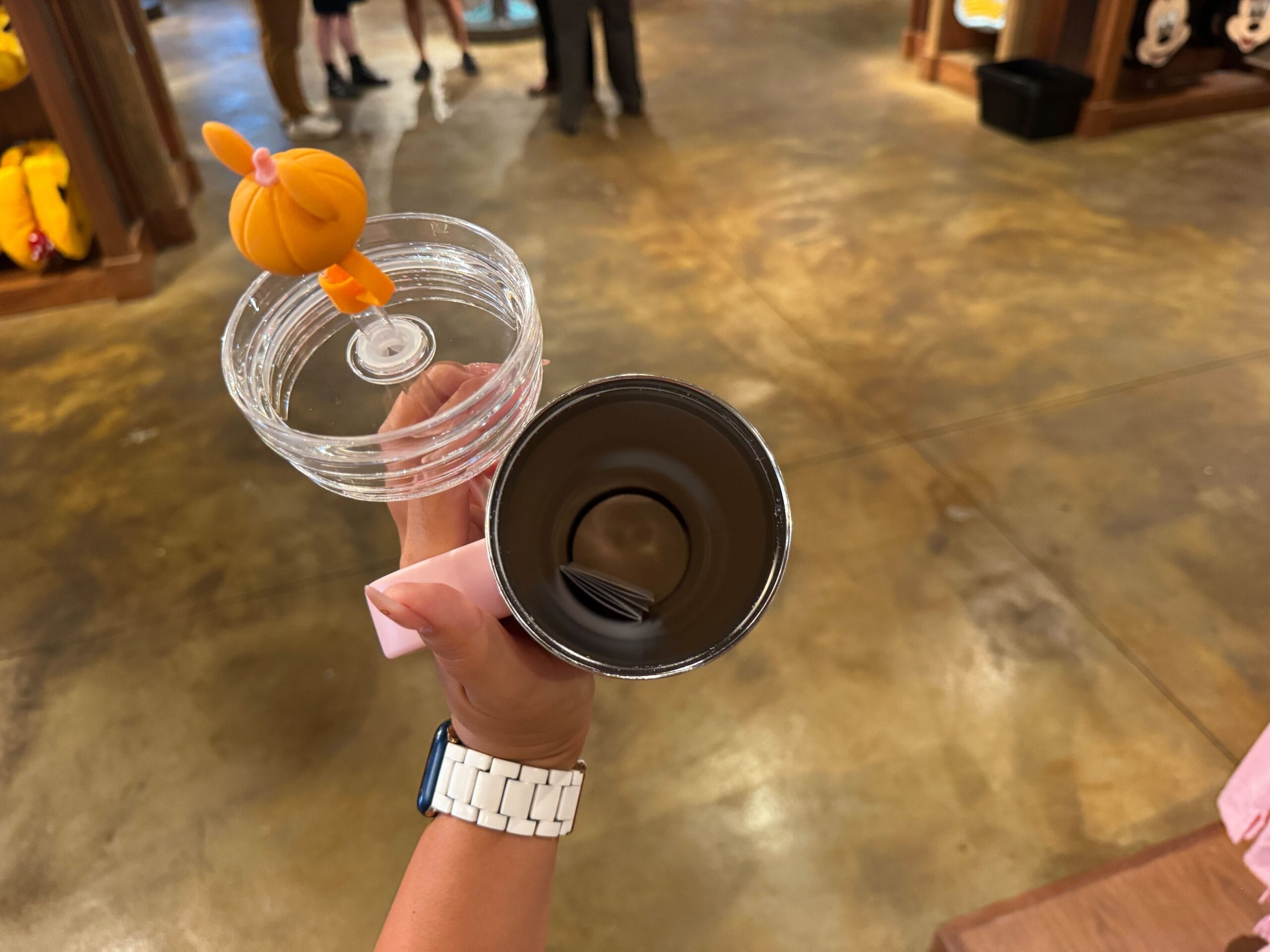 A hand holds an open tumbler with a clear lid featuring an orange Halloween merchandise figure on top in a store.