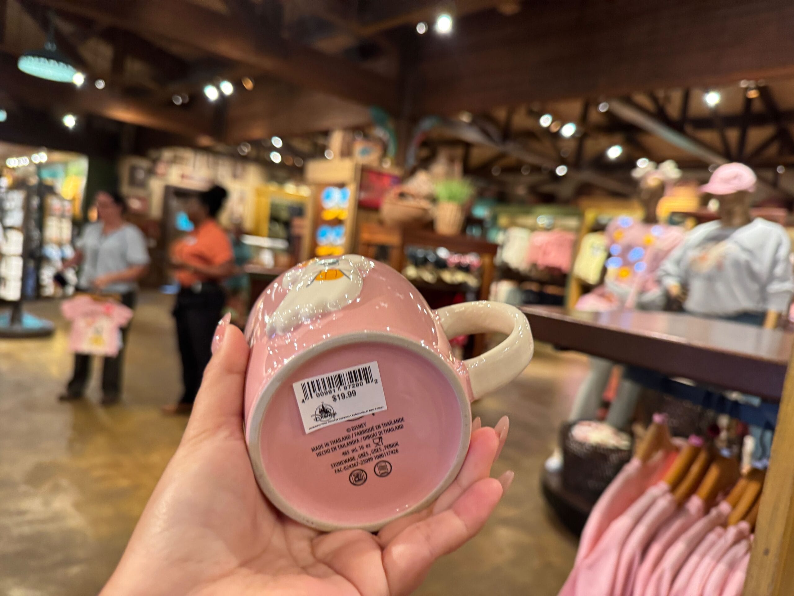 A hand holds a pink mug with a barcode—Halloween merchandise at Walt Disney World, shoppers and clothing in view.