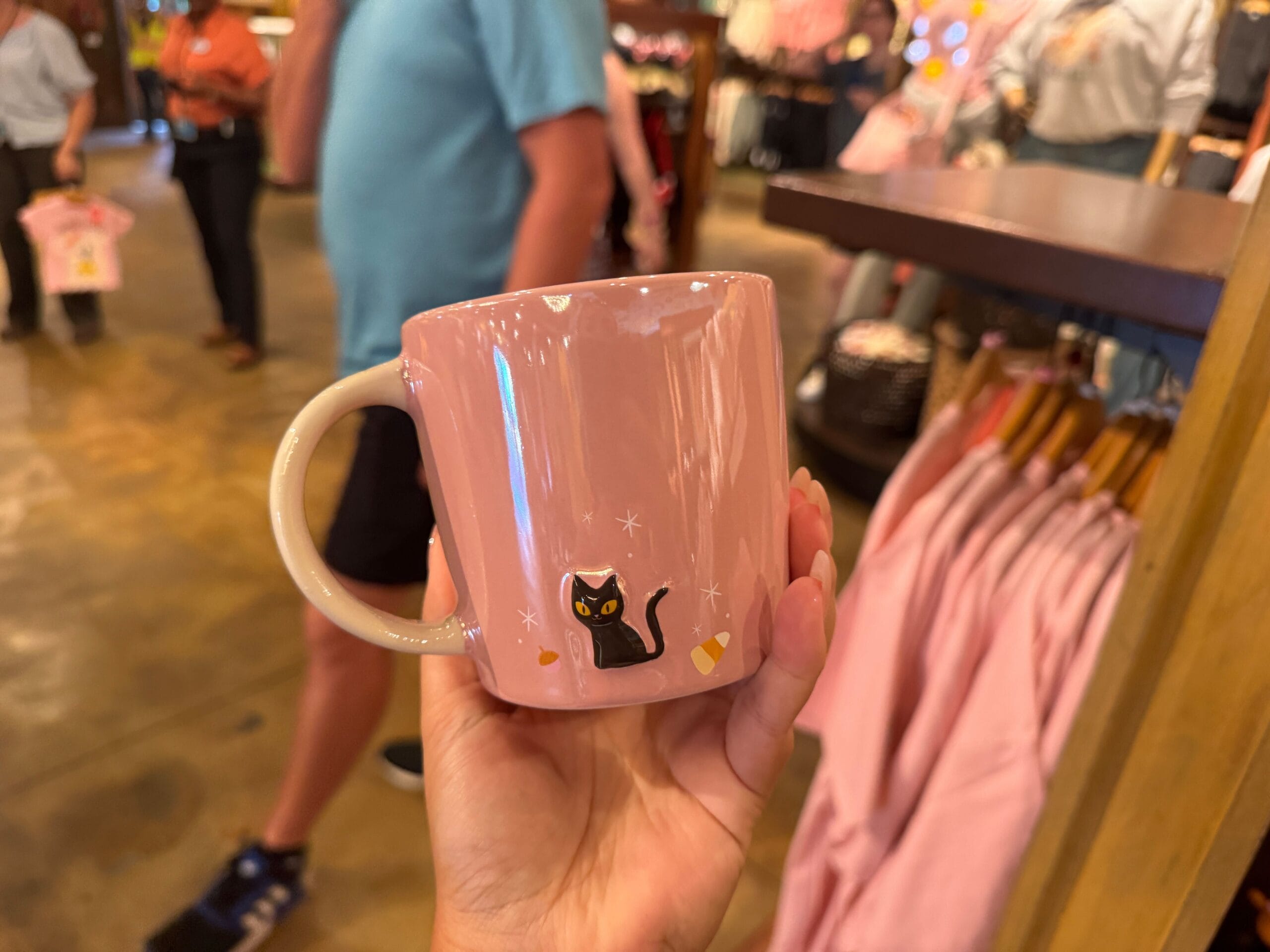 A hand holds a pink mug with a black cat design, part of the Halloween merchandise at Walt Disney World.