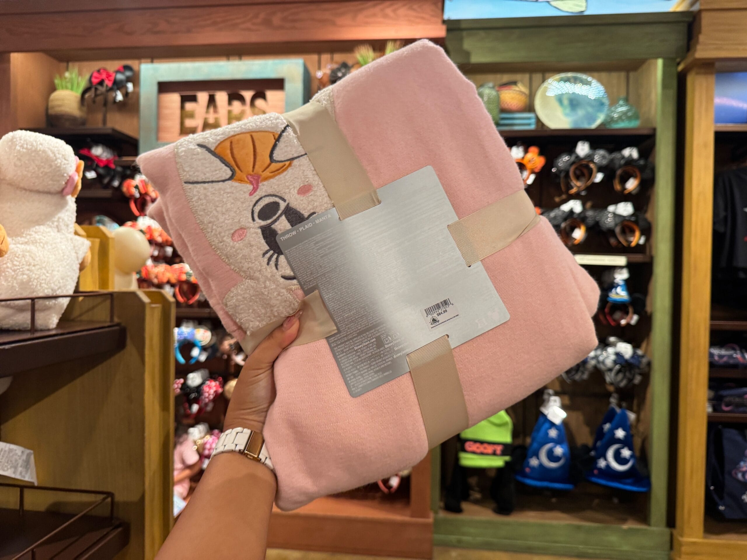 A hand holds a pink cat blanket among Halloween merchandise in a store filled with Walt Disney World items.