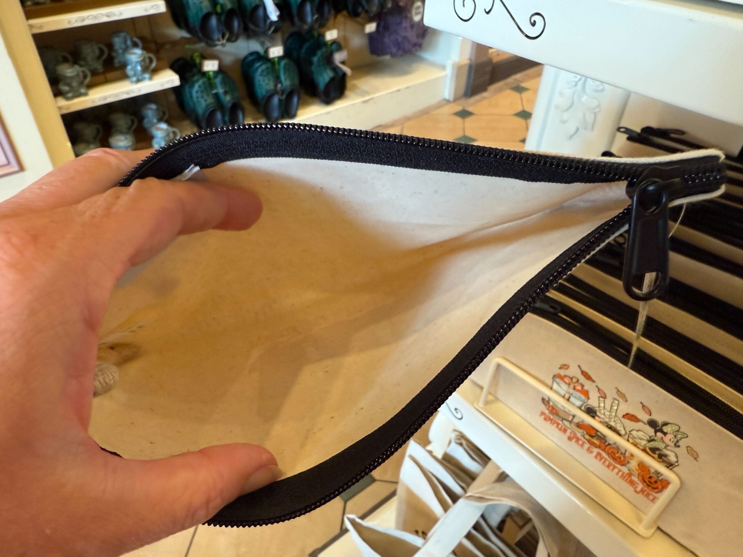 A hand holds open an empty beige zipper pouch in a store with Disney Halloween merch and souvenirs on nearby shelves.