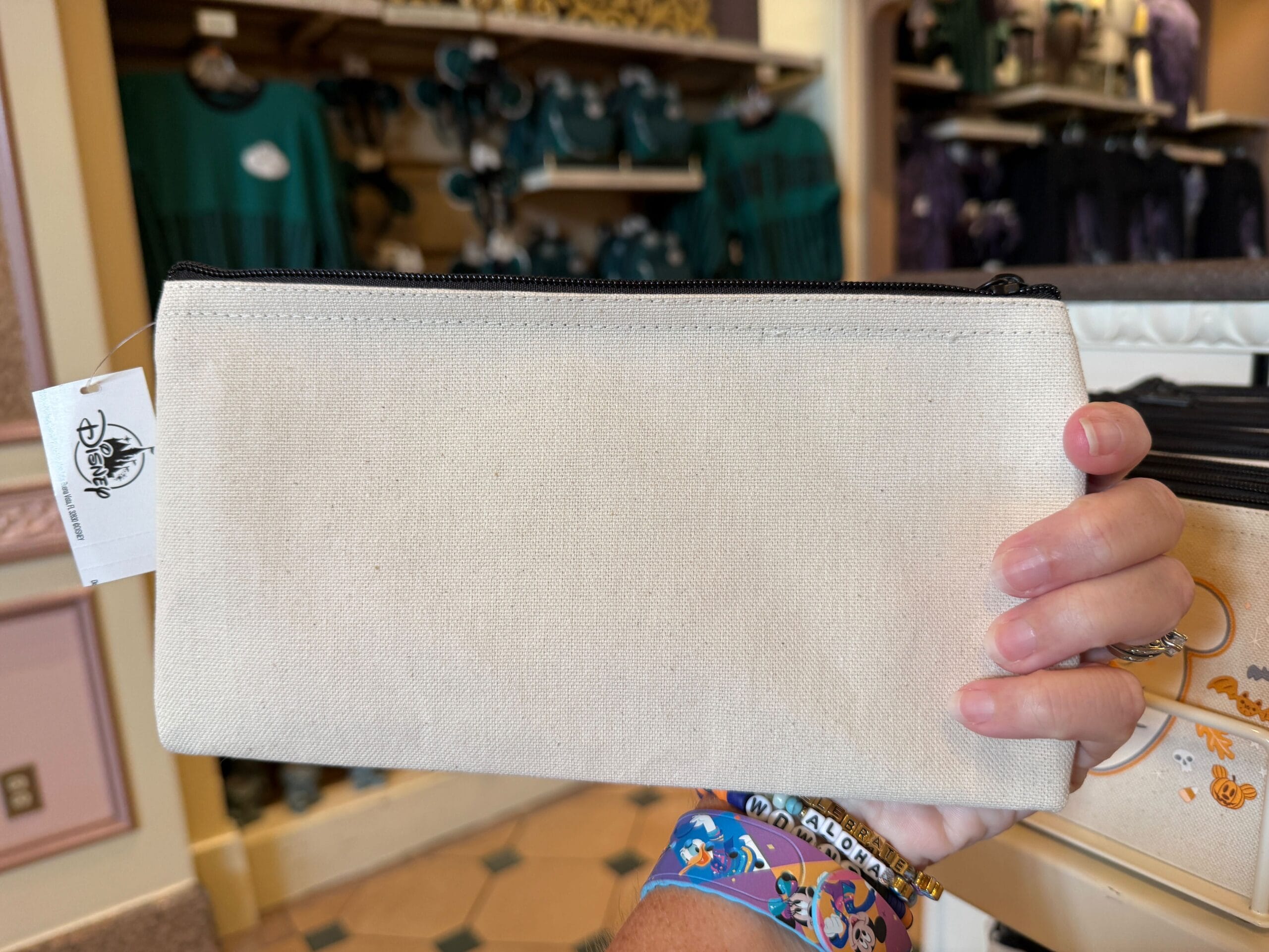 A hand holds a beige zippered pouch; Walt Disney World shelves with Disney Halloween Merch 2025 are blurred in the background.