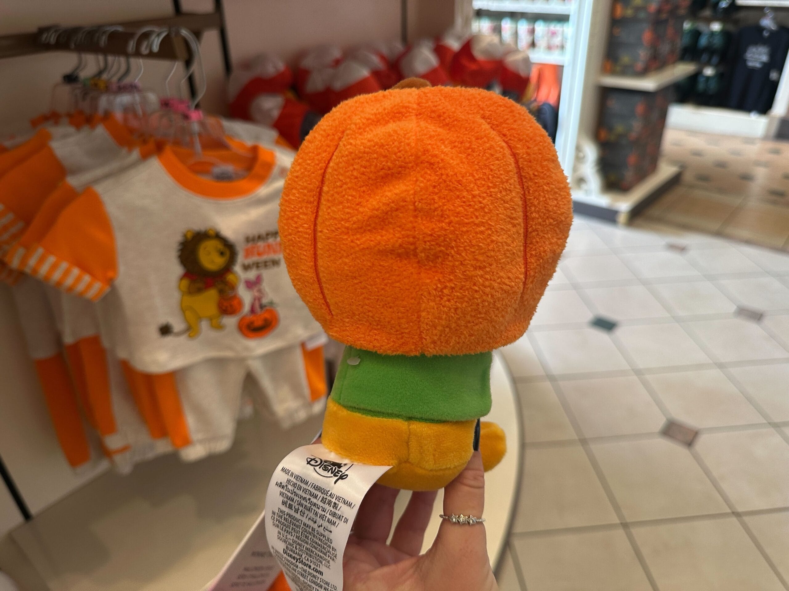 A hand holds a small plush toy dressed as a pumpkin among Disney Halloween Merch in a store with festive baby clothes.