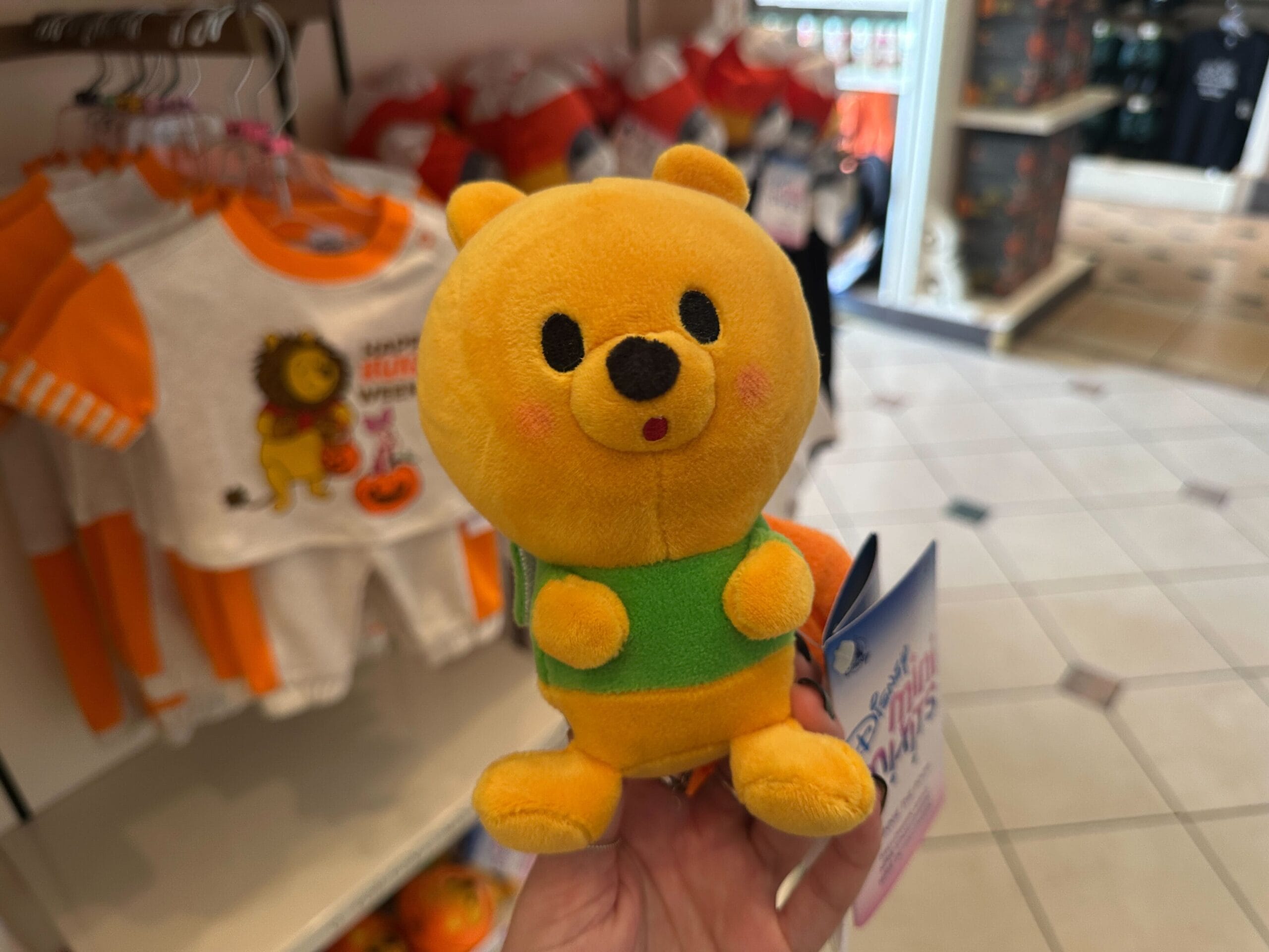 A hand holds a small plush bear in a green shirt, surrounded by Disney Halloween merch and baby toys in the background.