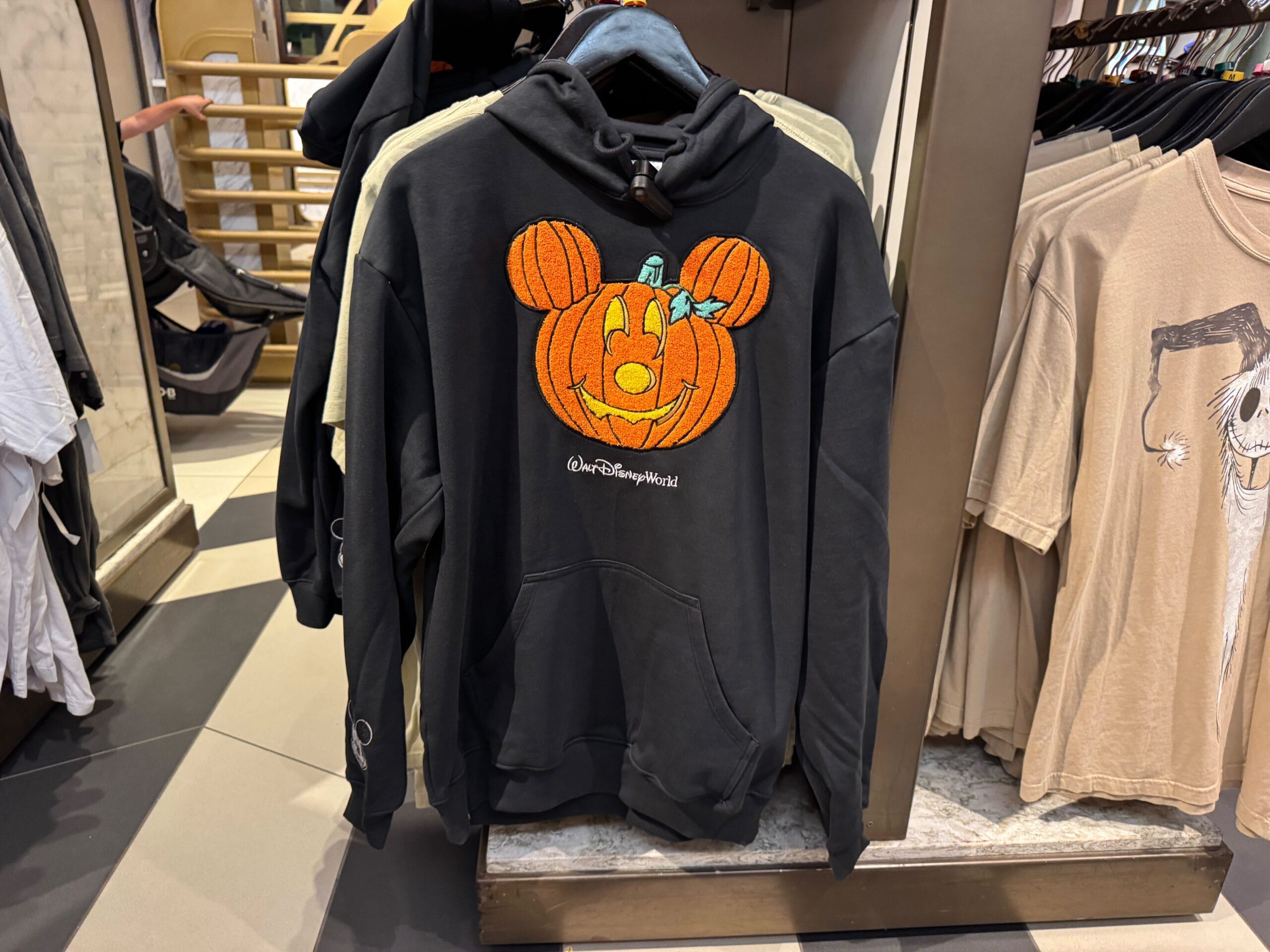 Black hoodie featuring a Mickey pumpkin design from 2025 Disney Halloween merch, spotted on a shop rack at a Disney park.