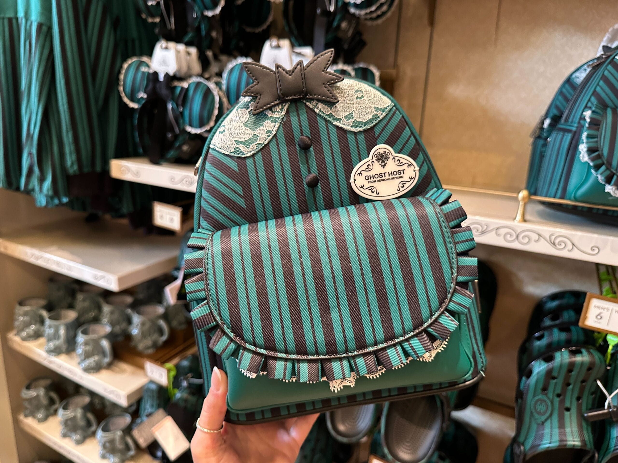 A hand holds a green and black striped mini backpack with ruffles, lace, and a “Ghost Host” name tag.