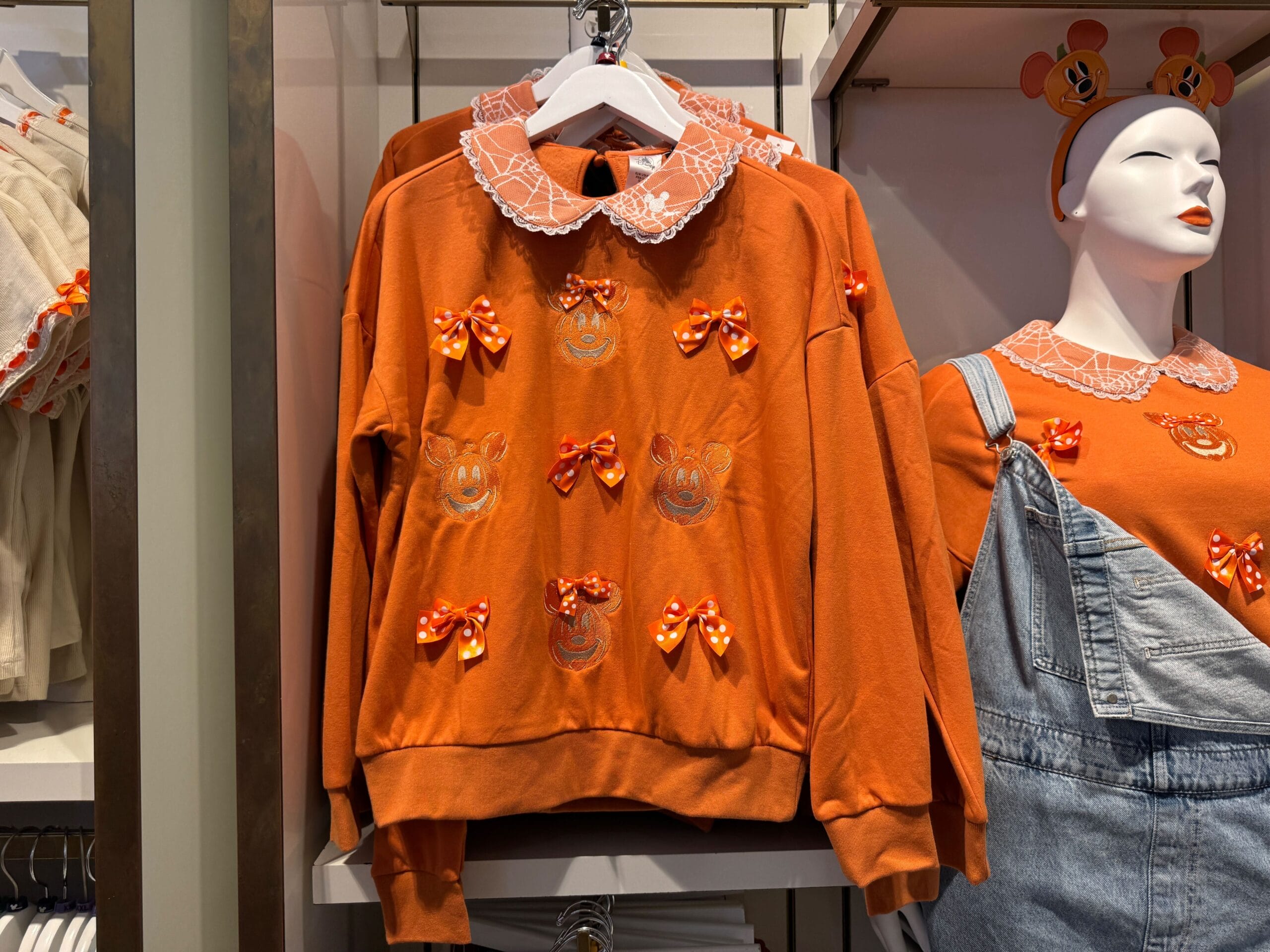 Orange sweatshirt with embroidered Mickey faces and bows, displayed on a hanger next to a mannequin in matching attire.