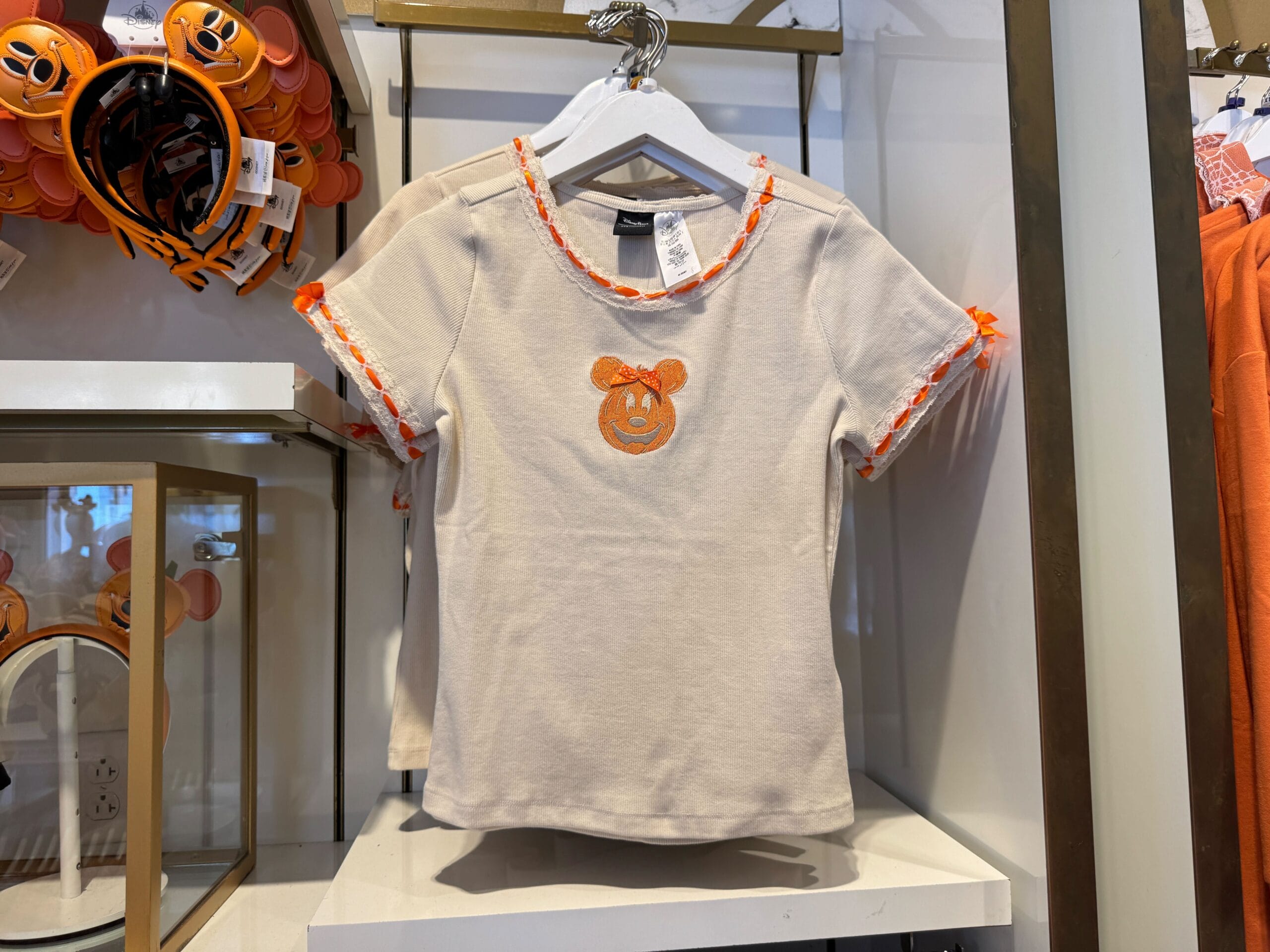Beige T-shirt with an orange Minnie Mouse pumpkin face displayed on a store rack with Halloween-themed accessories nearby.