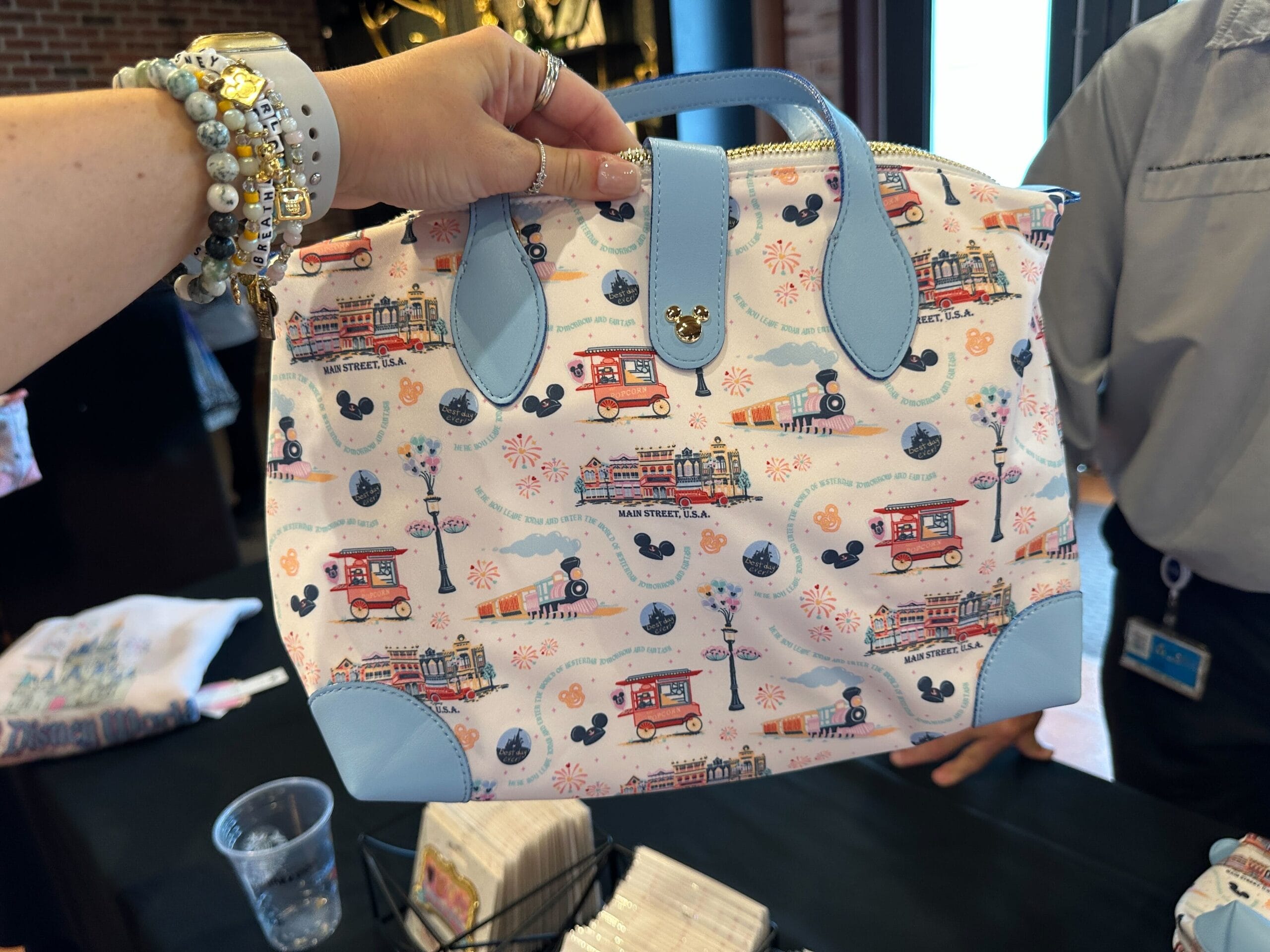 A hand holds a pastel Disney-themed handbag decorated with Main Street, U.S.A. icons and blue handles.