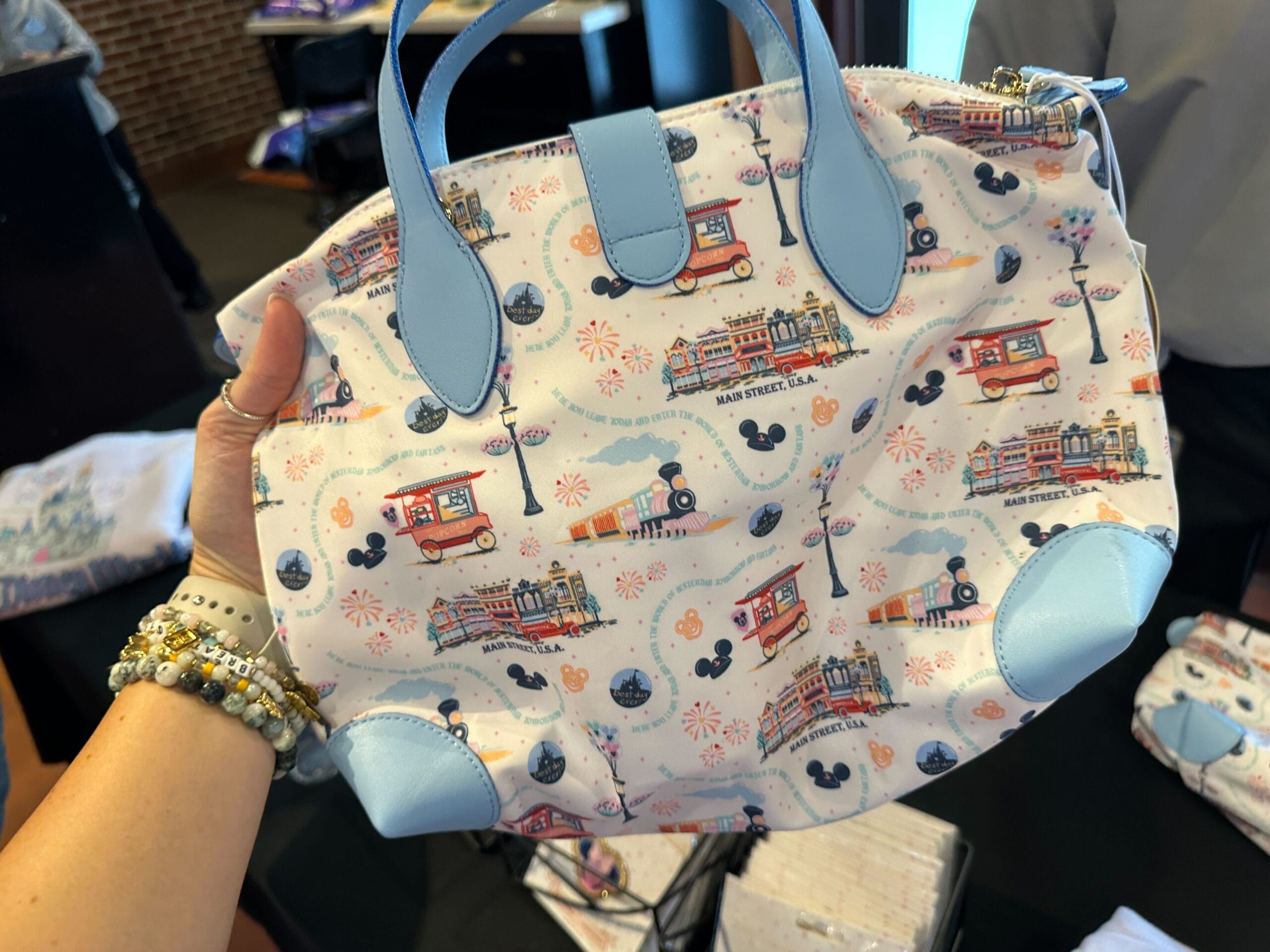 Hand holding a pastel purse with Disney-themed prints of Main Street vehicles, Mickey icons, and fireworks.
