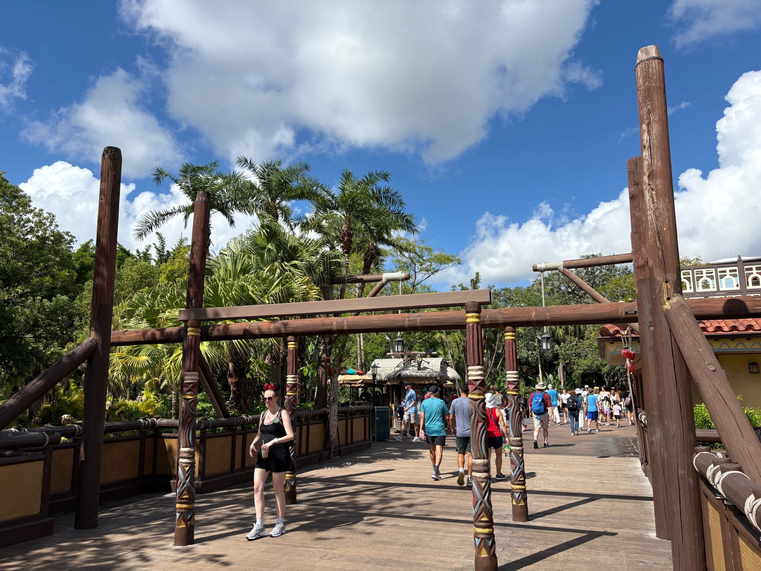 People walk on a wooden bridge surrounded by tropical plants under a blue sky with scattered clouds.