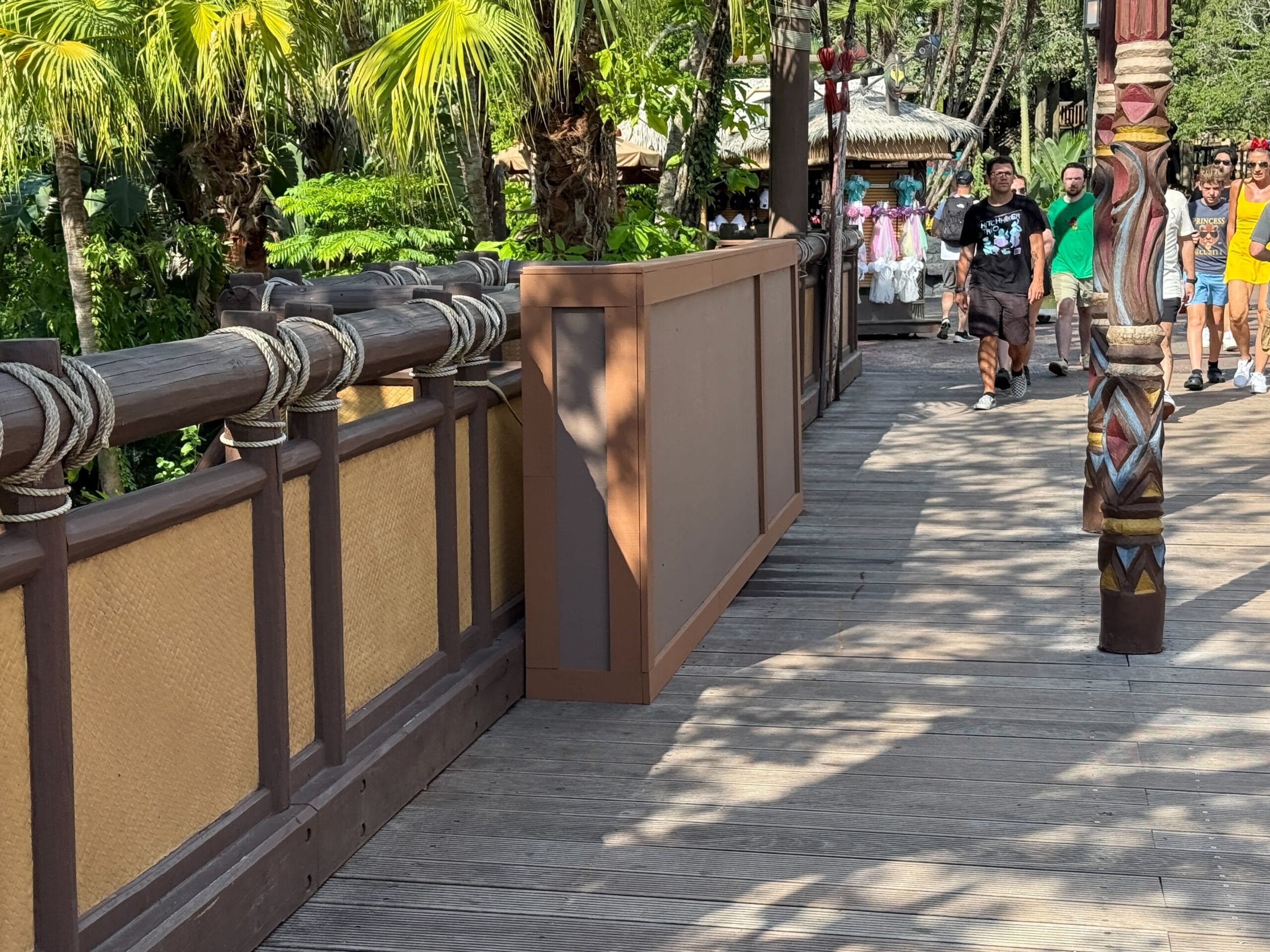 A wooden bridge with rope railings and tropical plants, with people walking in the background on a sunny day.