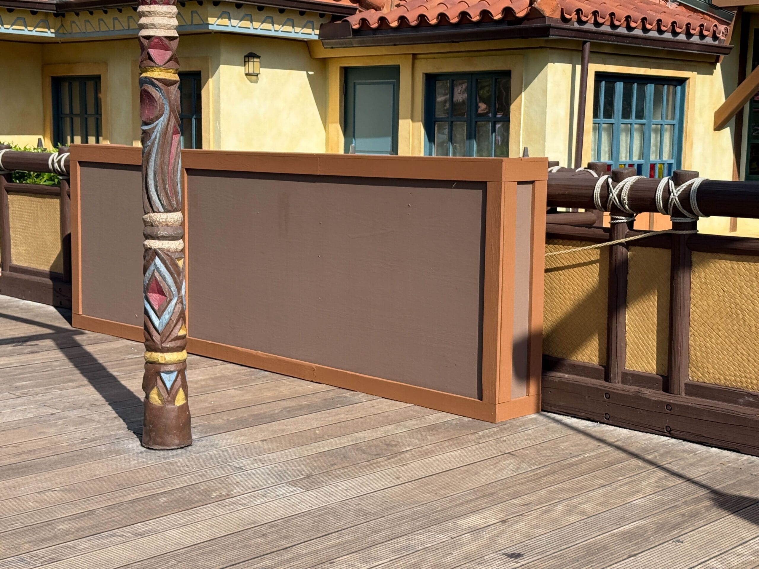 A wooden construction wall blocks part of a boardwalk next to a colorfully painted pole and tan building.