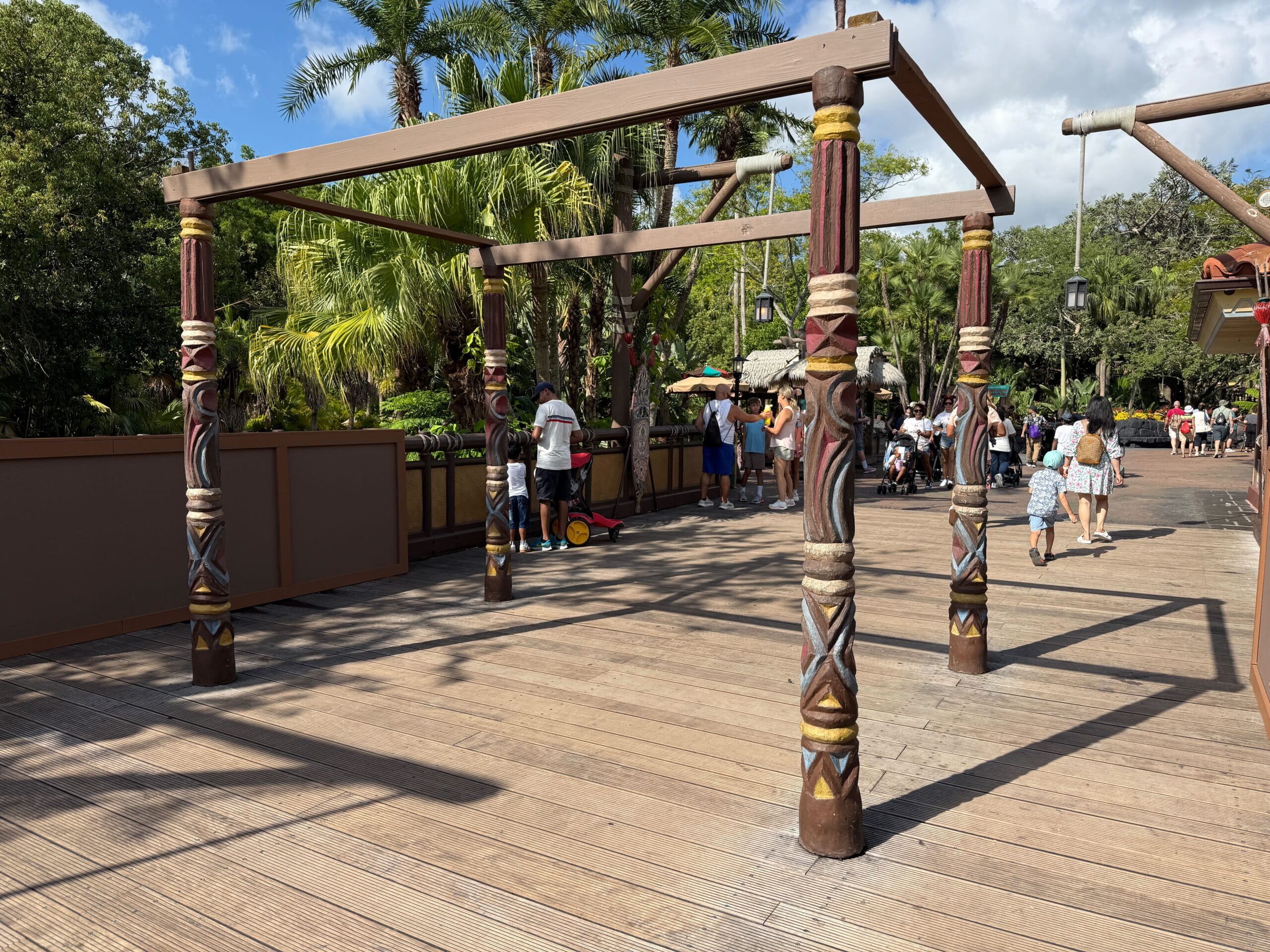 Wooden pergola with carved pillars in Adventureland, people relaxing under partly cloudy skies near the Adventureland sign.