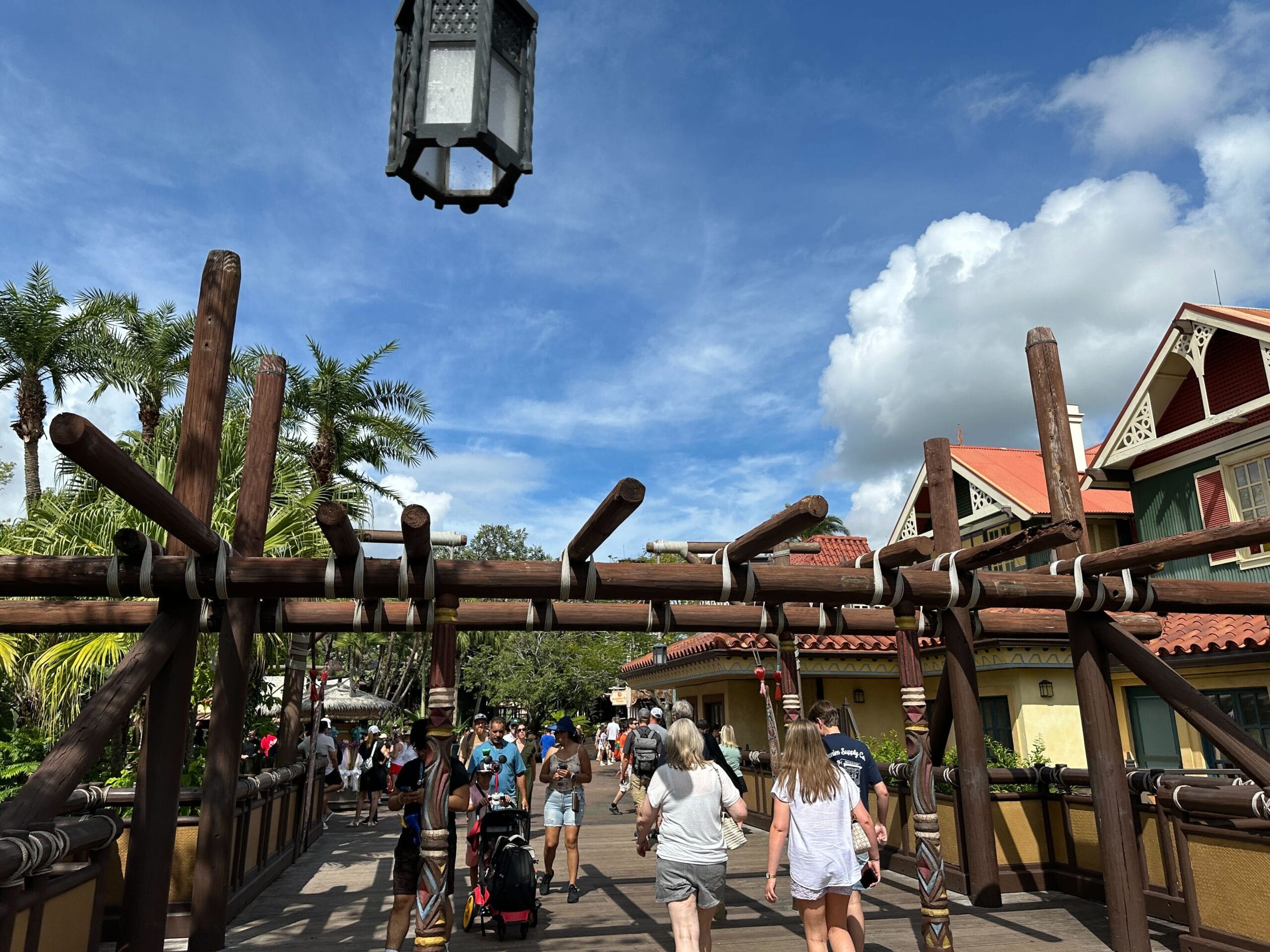 People walk under a wooden archway at a sunny outdoor location with palm trees and buildings in the background.