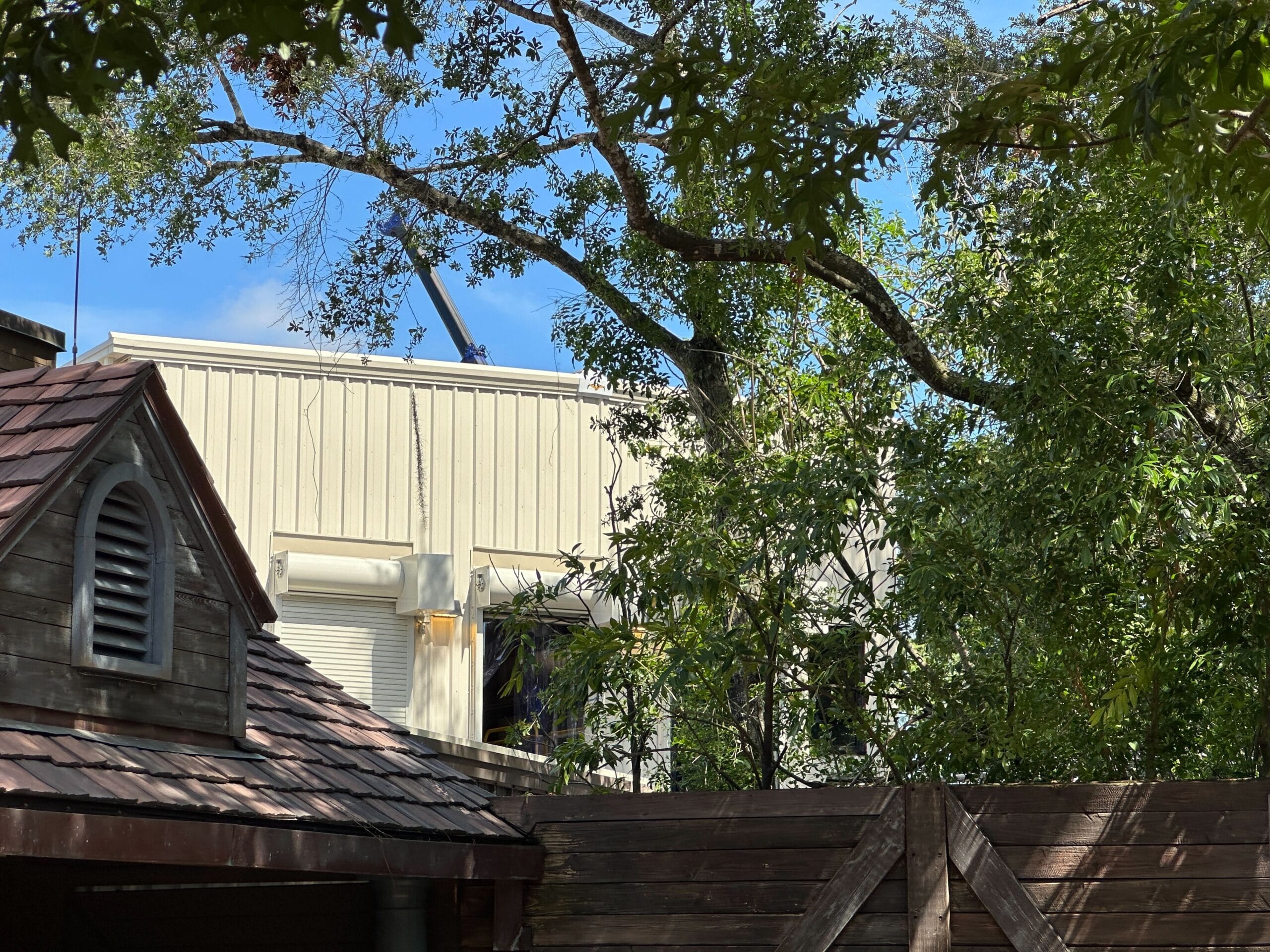 Wooden fence and roof in foreground, leafy trees, and a beige industrial building partially visible in the background.