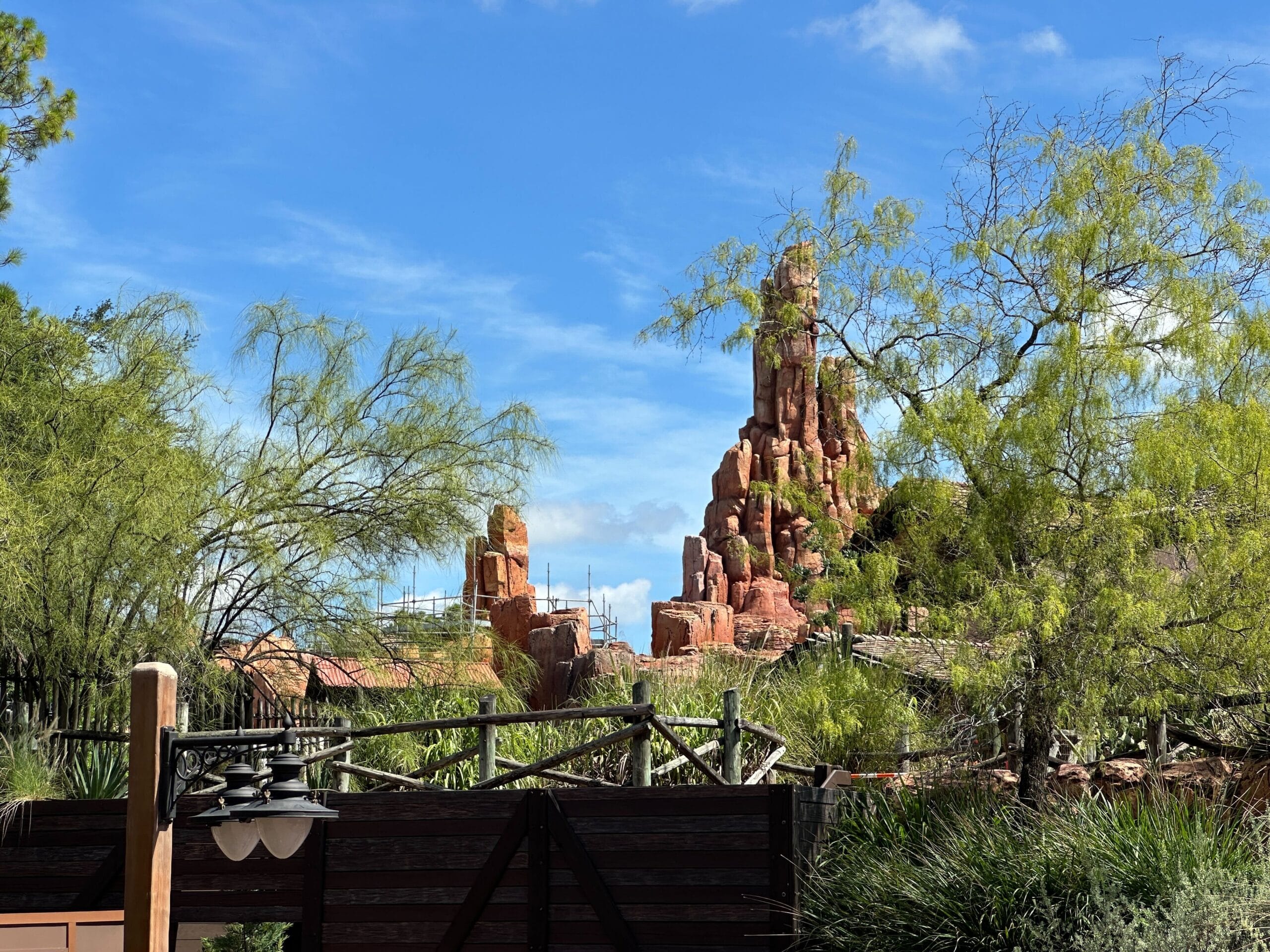 Red rock mountain structure behind trees and a wooden fence under a bright blue sky with scattered clouds.