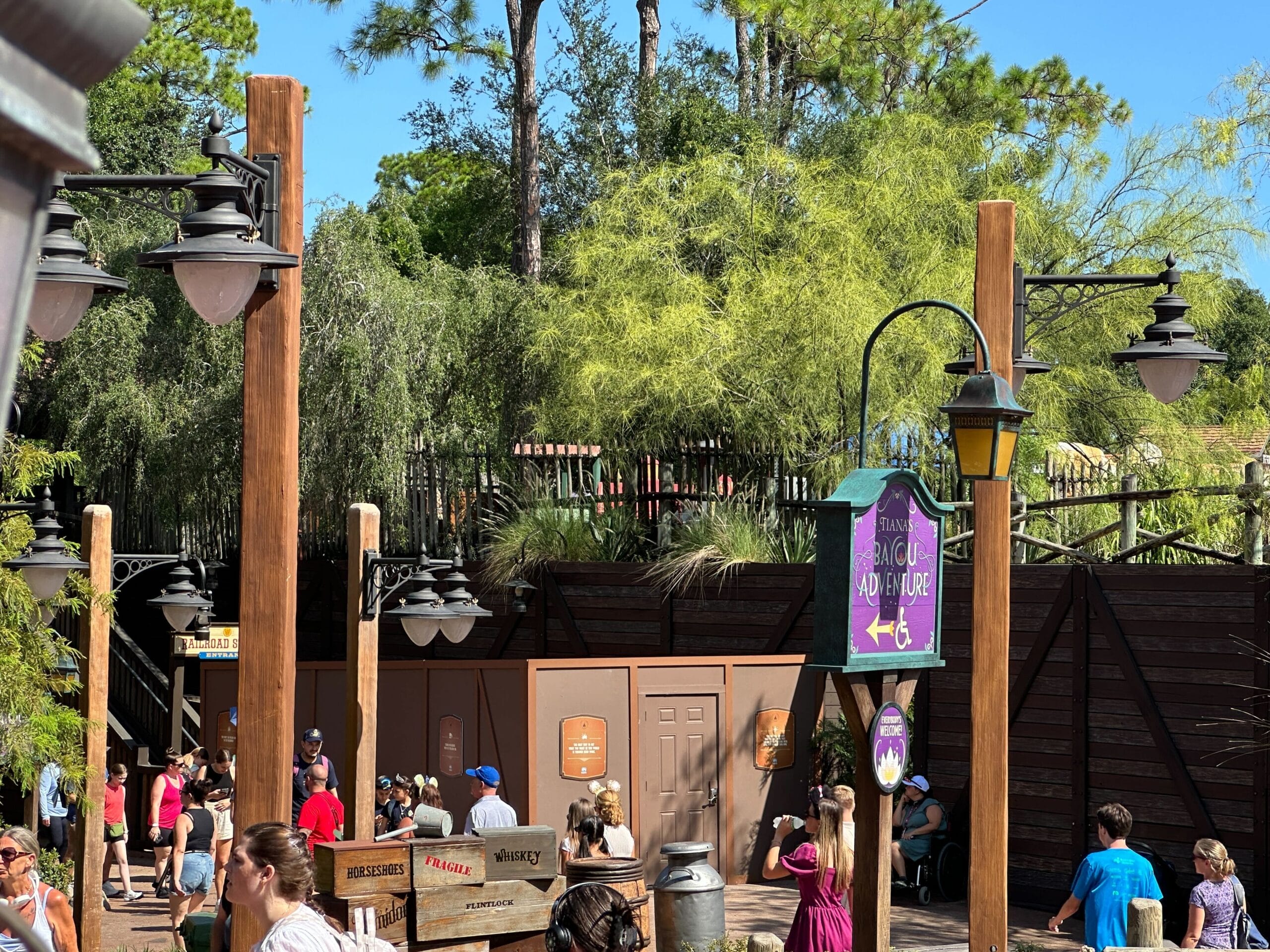 People walking and sitting near brown construction walls at an outdoor area with trees, lamps, and a cartoonish sign.