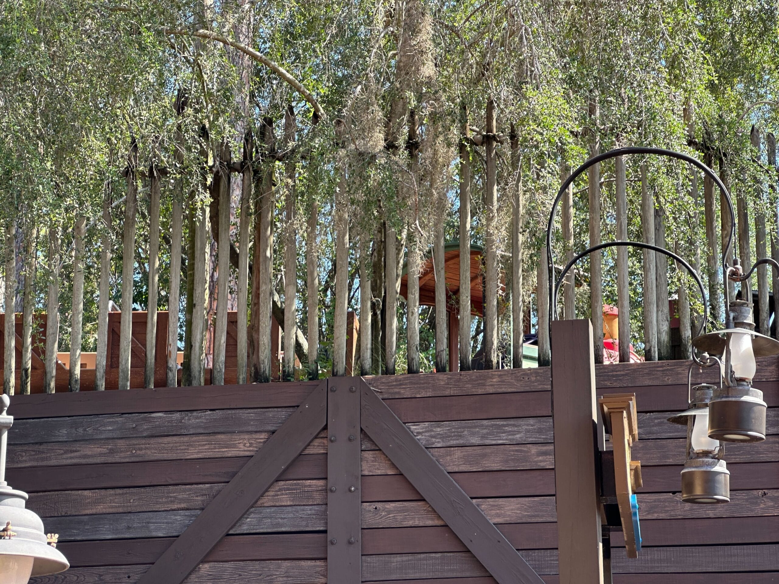 Wooden fence with trees behind it, hanging lanterns, and a round table partially visible in the background.