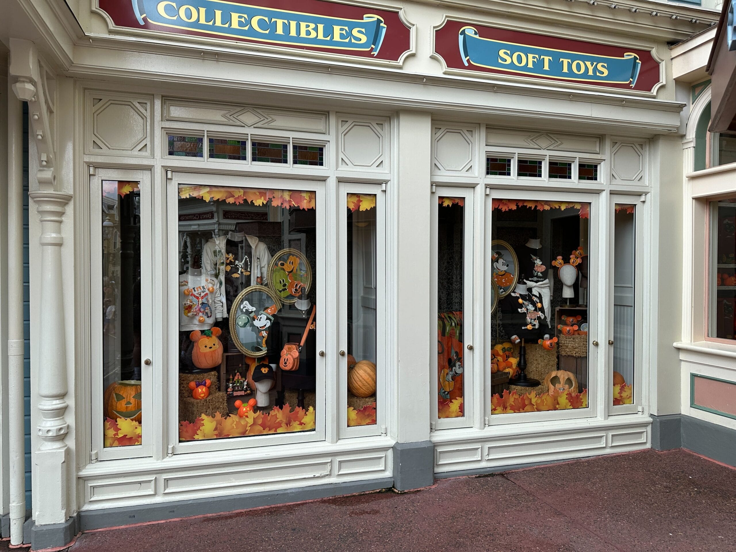 A shop window decorated with pumpkins and autumn leaves for Halloween, featuring plush toys and collectibles inside.