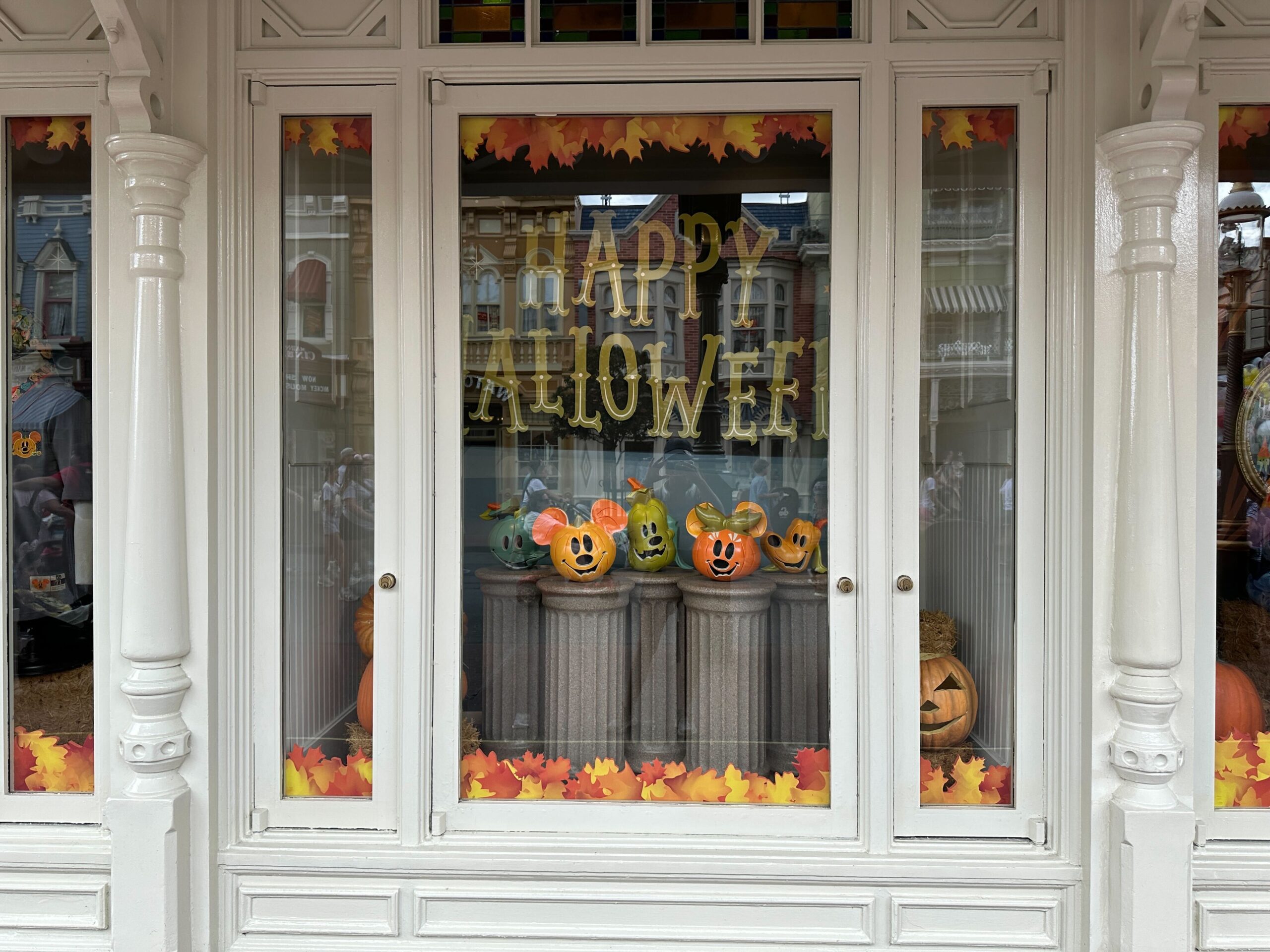Storefront window with "Happy Halloween" sign and colorful painted pumpkins displayed, decorated with autumn leaves.