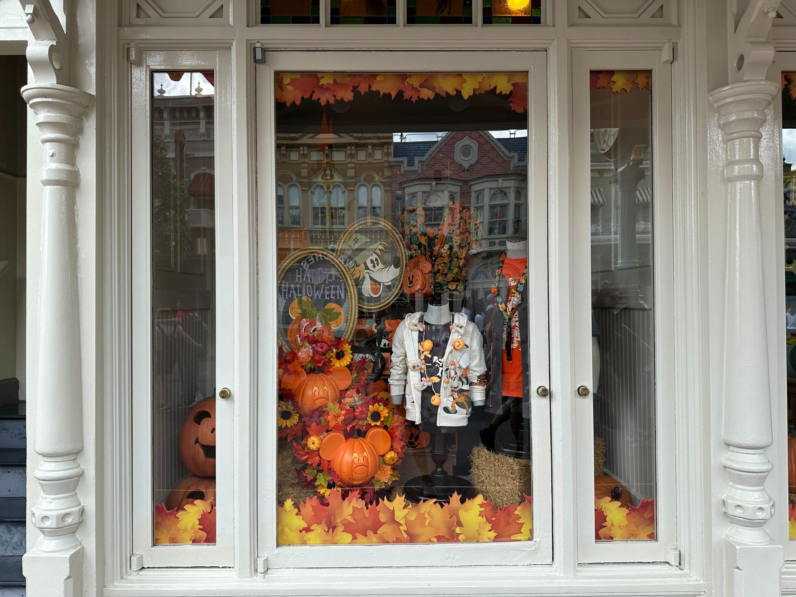 Store window decorated for Halloween with pumpkins, autumn leaves, and festive clothing on mannequins.