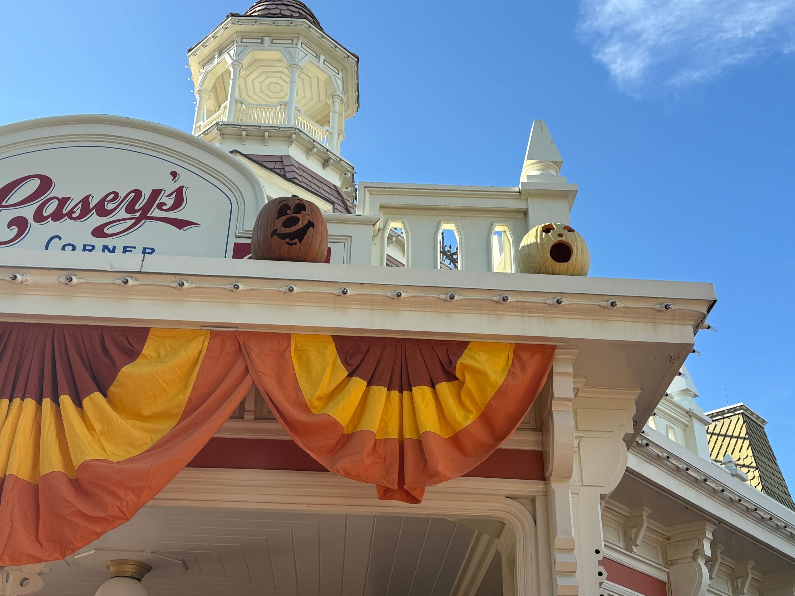 Two Jack-o'-Lanterns sit atop a building with festive Halloween decor and bunting under a bright blue sky.