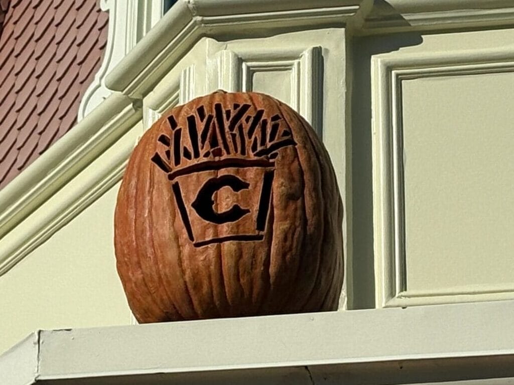 A carved pumpkin "C" Jack-o'-Lantern decorates a building ledge under the blue sky at Magic Kingdom.