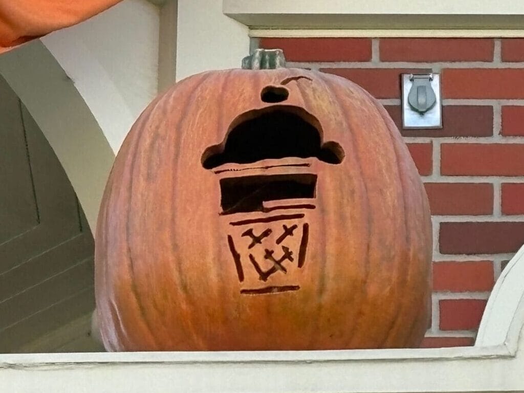 A Jack-o'-Lantern carved like a trash can sits on a ledge of a decorative, Magic Kingdom-inspired building.