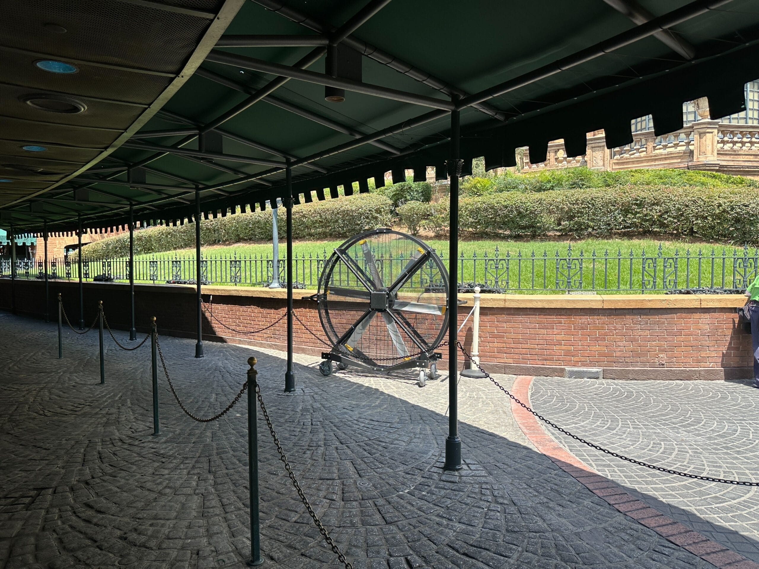 A shaded queue area with chains, a large fan, and a grassy hill with a brick wall and fence in the background.
