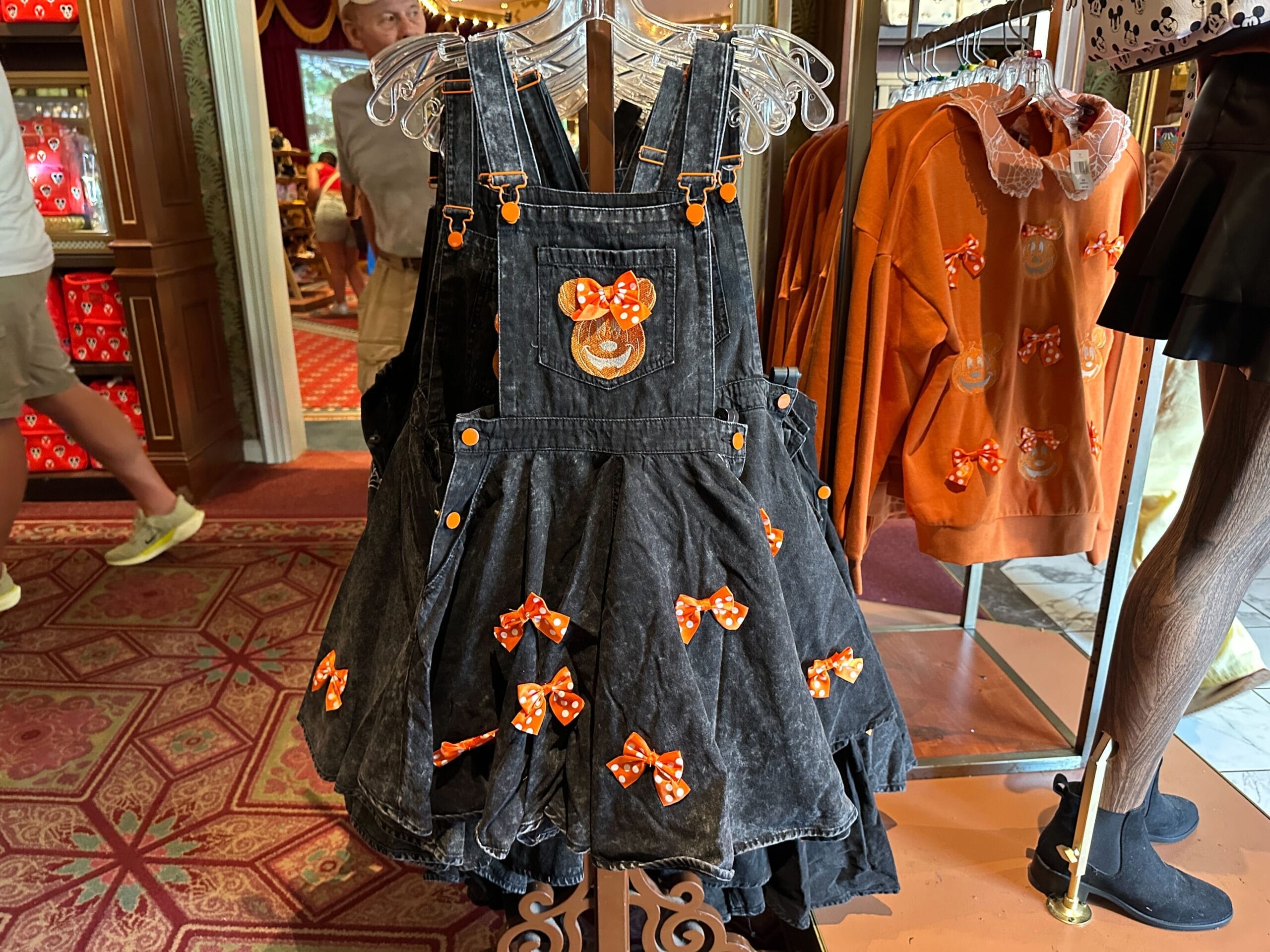 Black denim dress with orange Halloween Minnie Mouse bows by Her Universe, hanging on a rack in a clothing store.