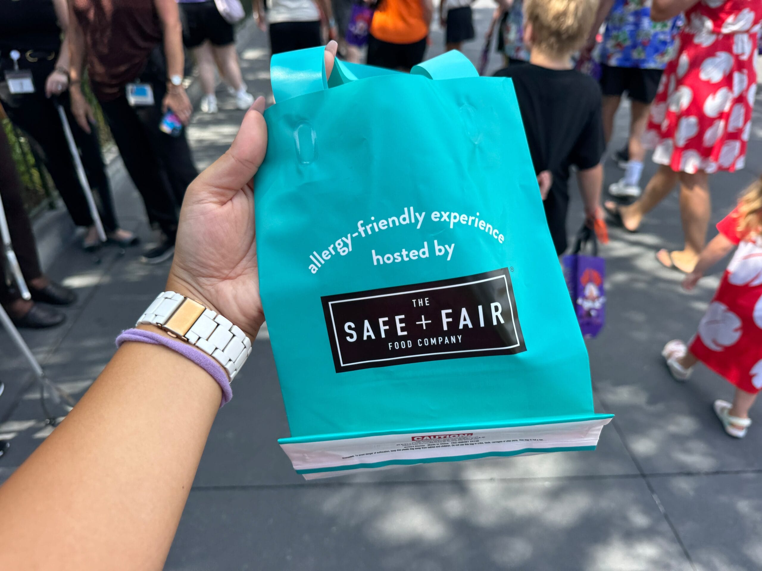 Hand holding a blue allergy-friendly treat bag from The Safe + Fair Food Company at a 2025 outdoor event with people in background.