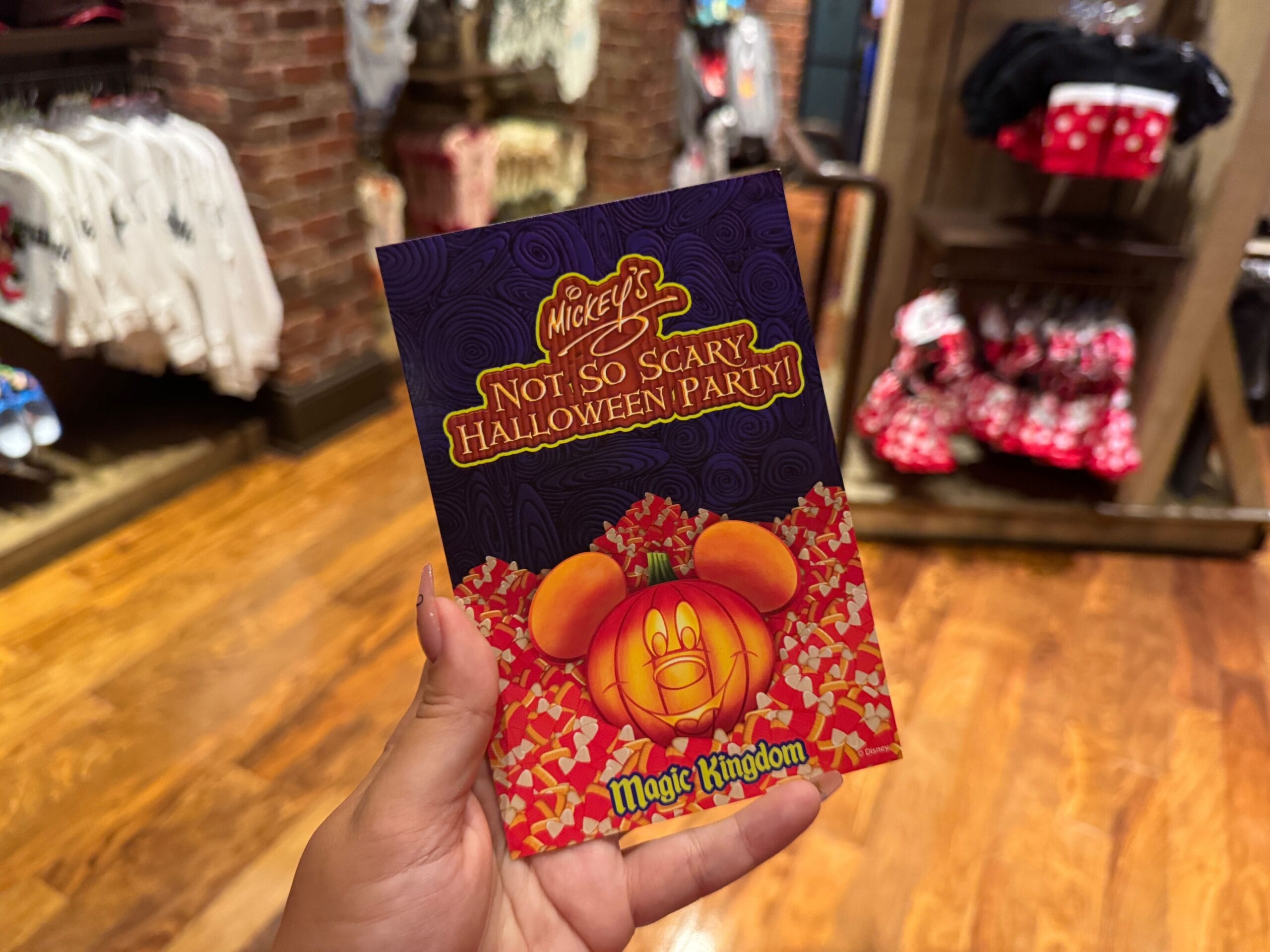 Hand holding a 2025 Mickey's Not So Scary Halloween Party signature card in a Disney gift shop, with character appearances nearby.