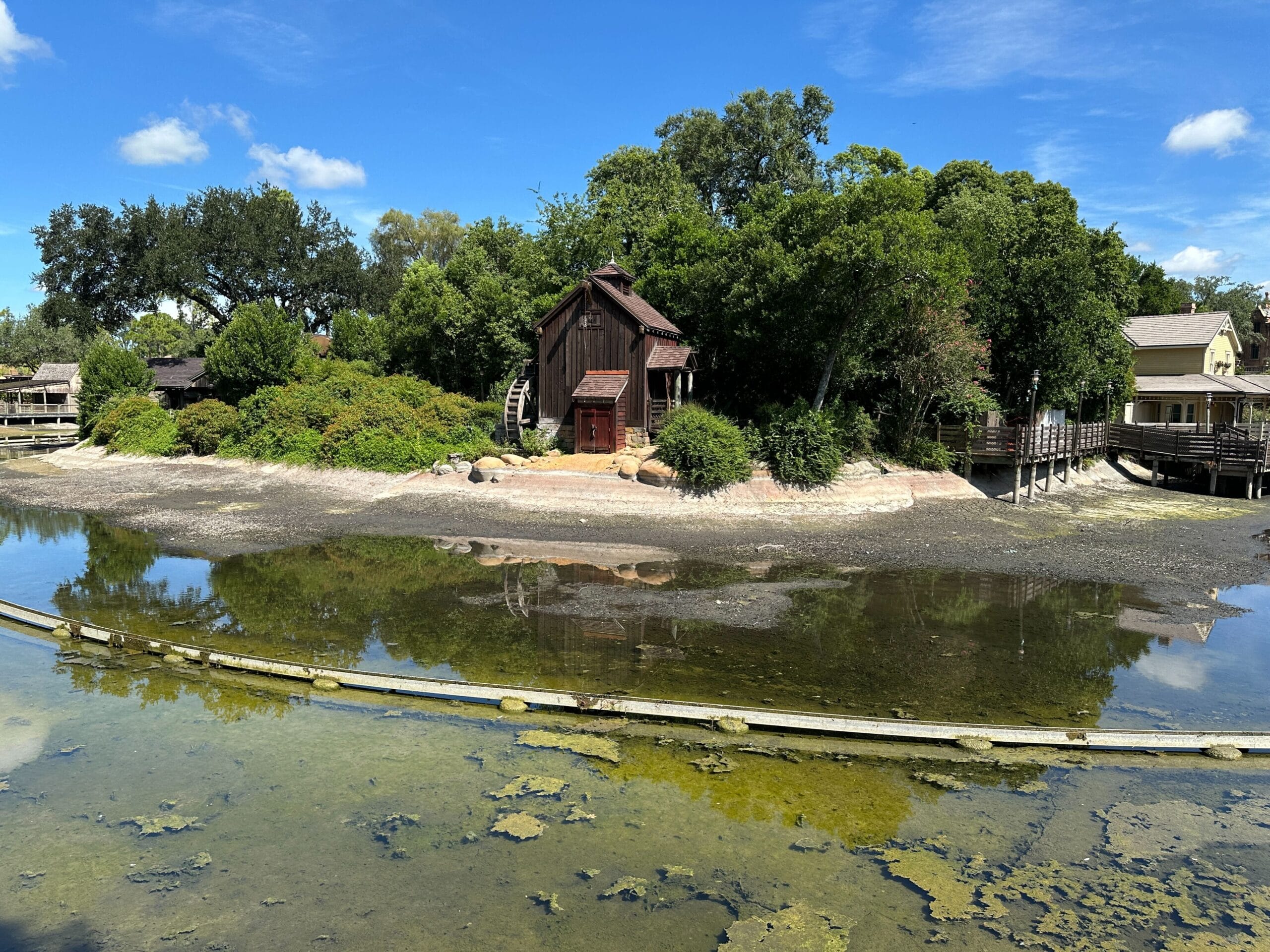 A rustic wooden cabin stands by a shallow, reflective pond surrounded by trees and greenery under a blue sky.